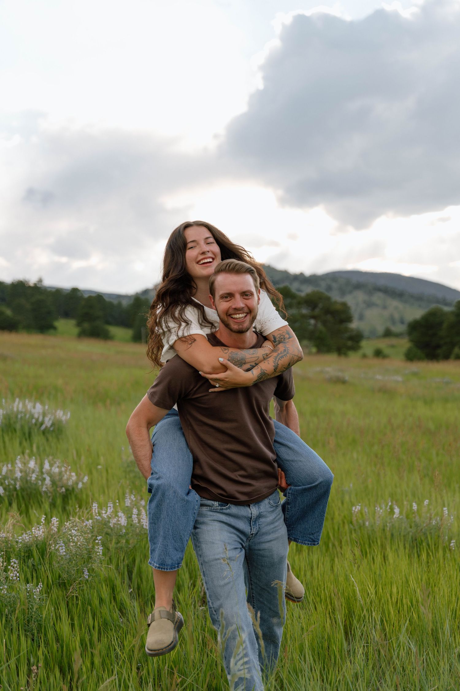 A playful couple enjoying a piggyback ride in a green meadow with mountains in the background.