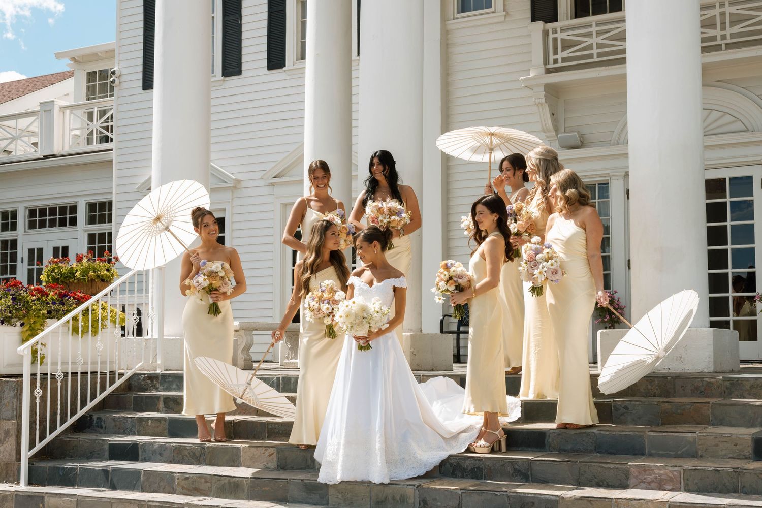 Bride in white gown with bridesmaids in champagne dresses holding parasols on steps of elegant white colonial venue.