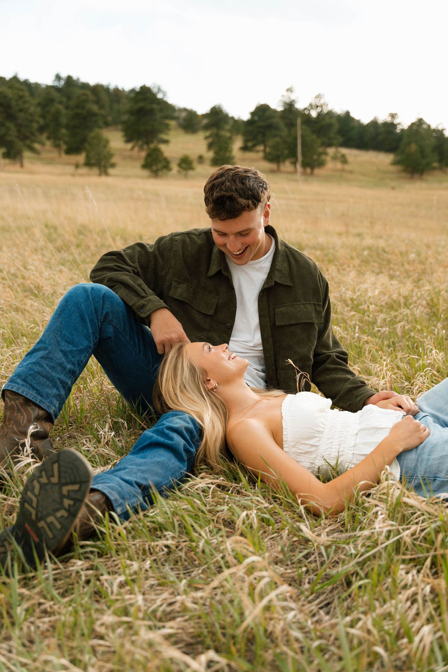 A couple relaxes in a golden meadow, woman lying in man's lap, both smiling, with pine-covered hills behind them.