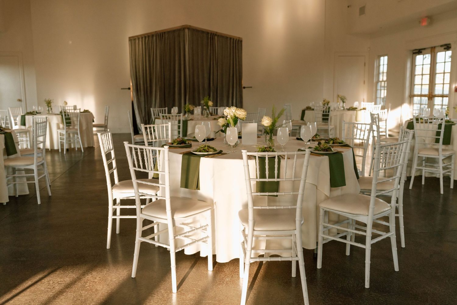 Elegant wedding reception hall with white Chiavari chairs, round tables with green and white linens, and floral centerpieces.