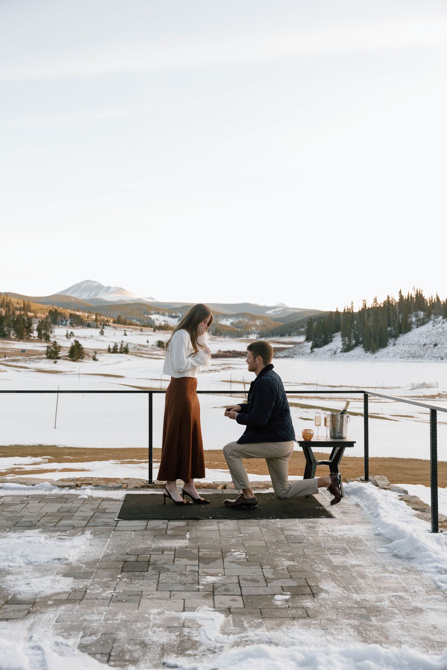 Marriage proposal on snowy mountain deck with champagne and scenic winter landscape.