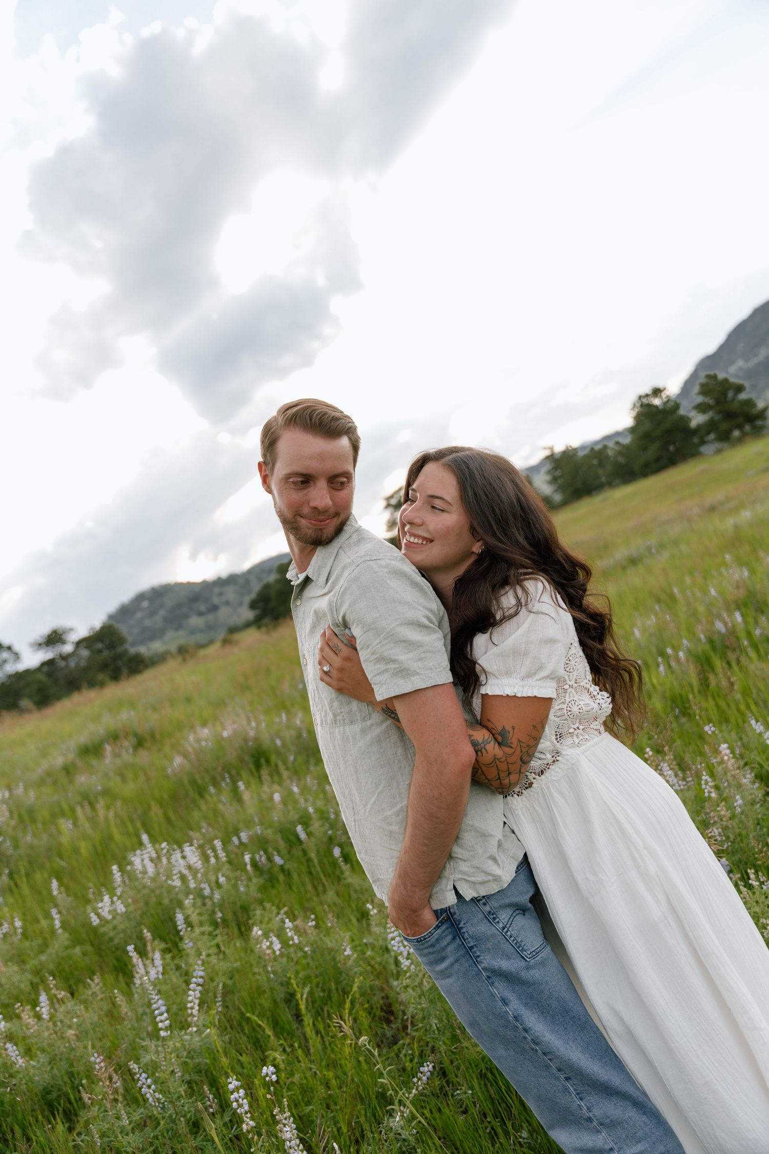A couple shares a playful piggyback ride in a mountain meadow filled with wildflowers.