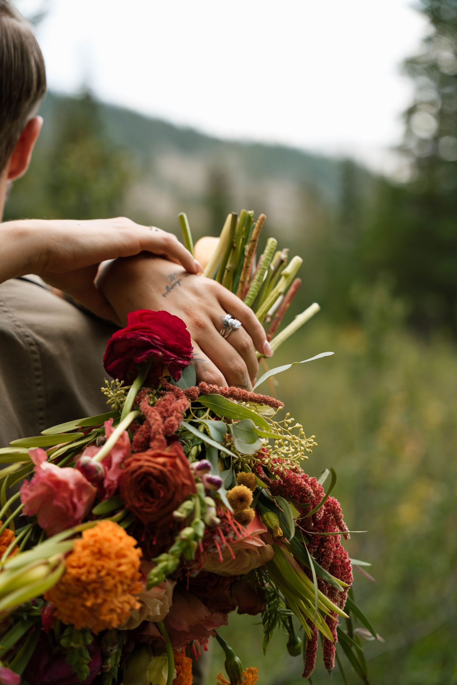 Person holding a lush bridal bouquet with red roses, marigolds, and greenery outdoors with mountain backdrop.