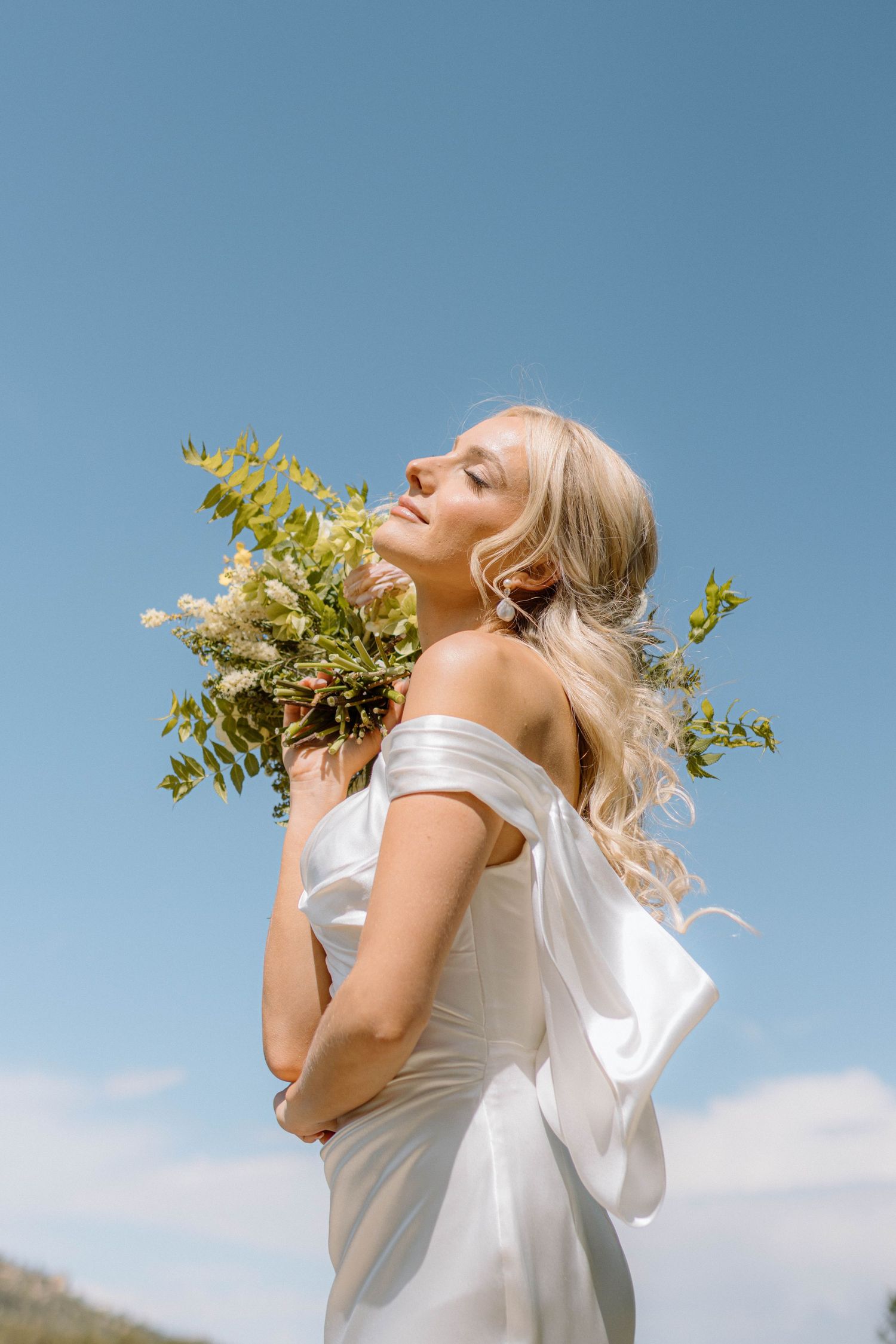 Bride in white off-shoulder gown holds wildflower bouquet against blue sky, eyes closed, blonde hair flowing.