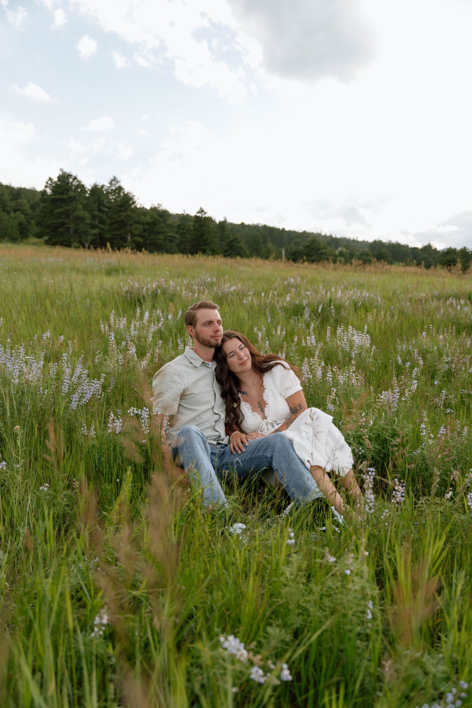 Couple sits together in tall grass and wildflowers with mountain landscape behind them.