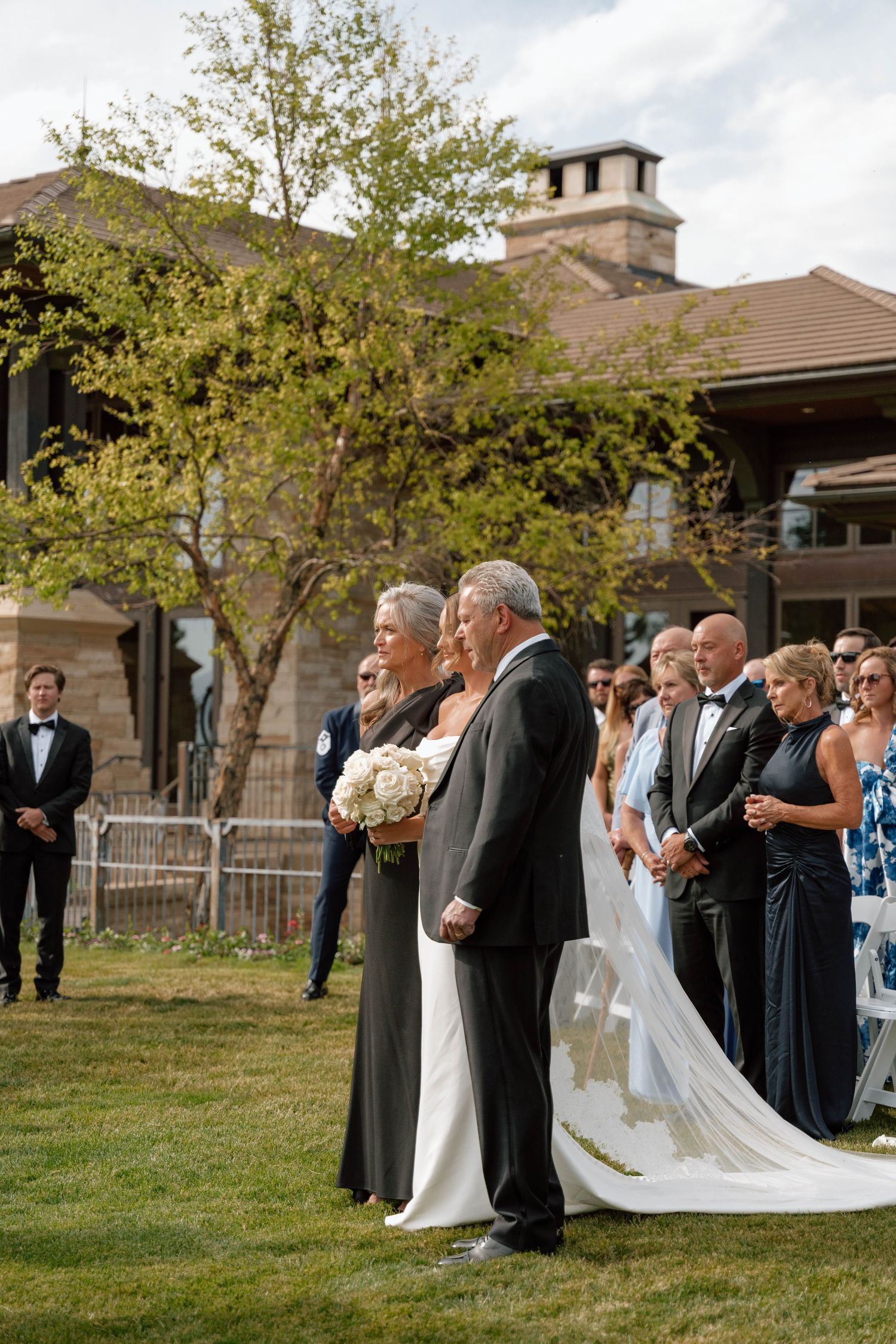 Elegant outdoor wedding ceremony in front of rustic stone building with wedding party in formal attire.