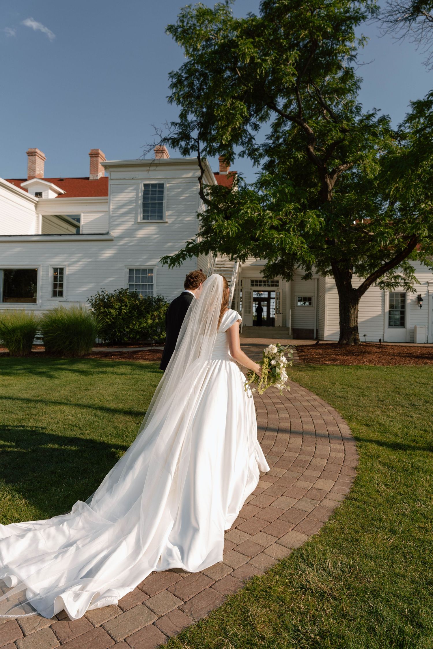 A bride in a flowing white gown walks along a curved brick pathway near a historic white house.