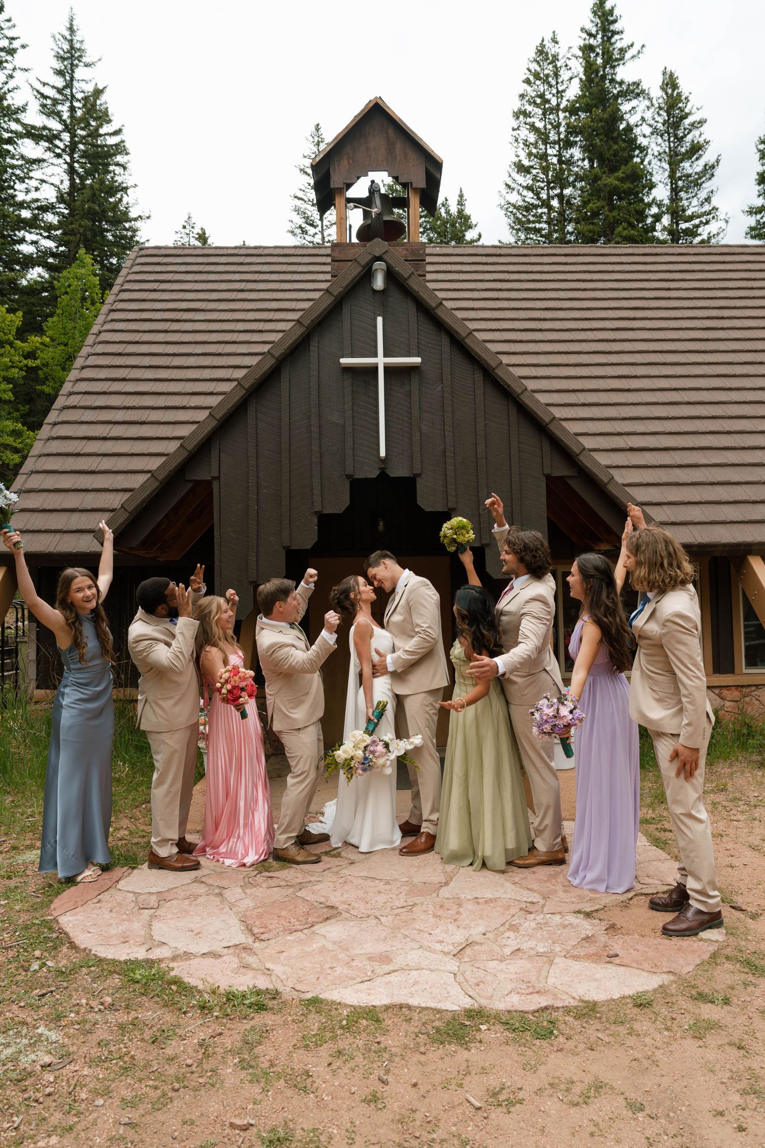 Wedding party celebrates outside a rustic wood chapel with a cross as the bride and groom share a kiss.