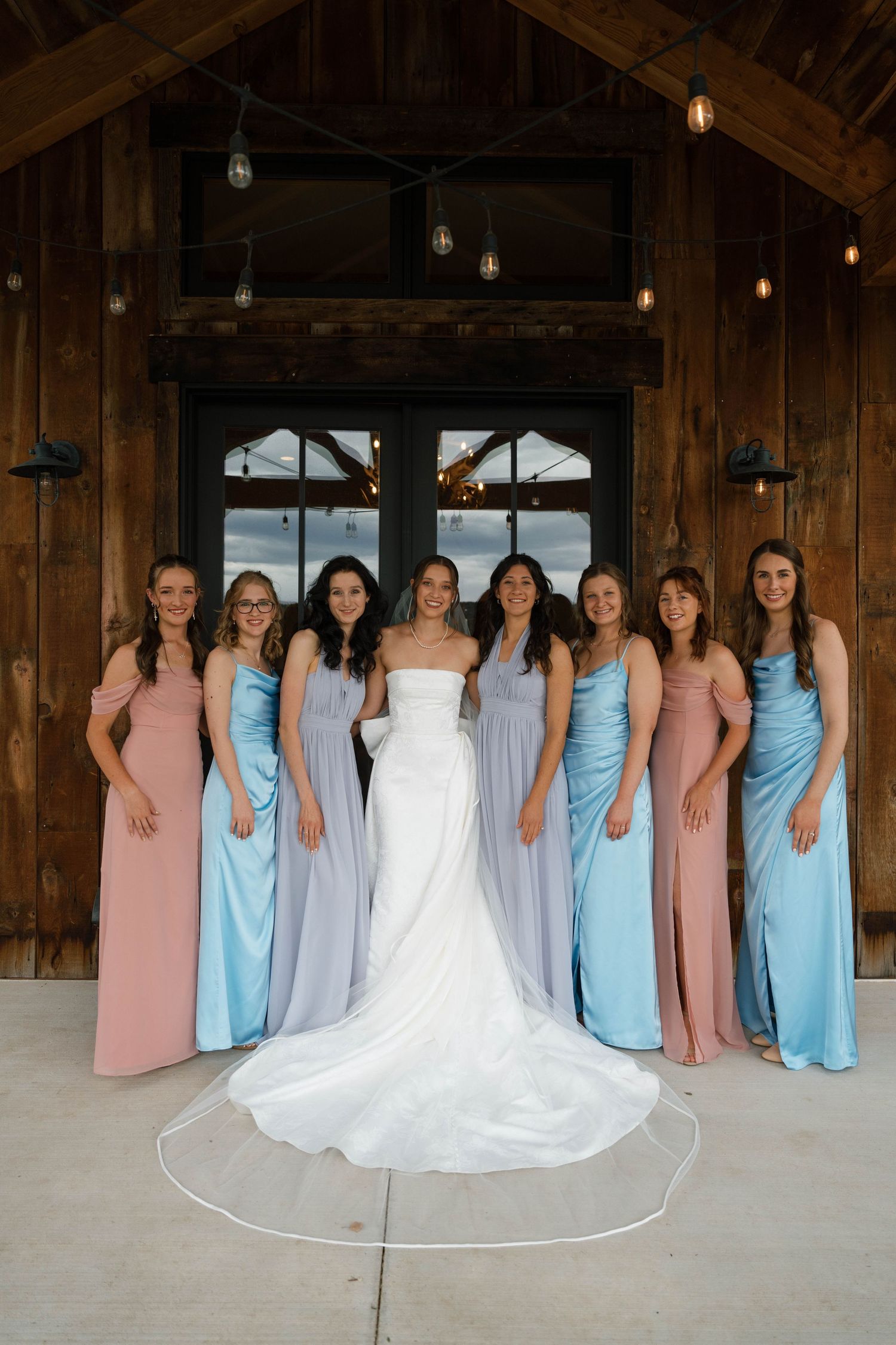 Bride in white gown poses with seven bridesmaids in pastel blue, lavender, and pink dresses inside a rustic barn venue.