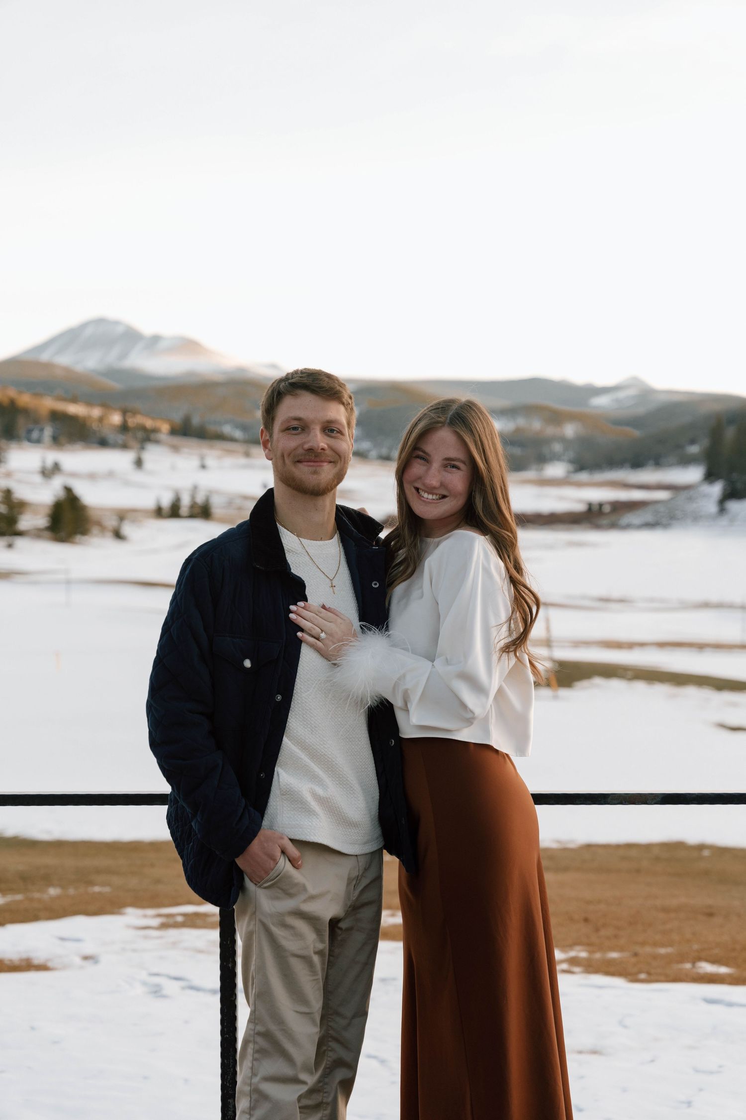 Engaged couple showing ring on mountain deck with snowy winter landscape.