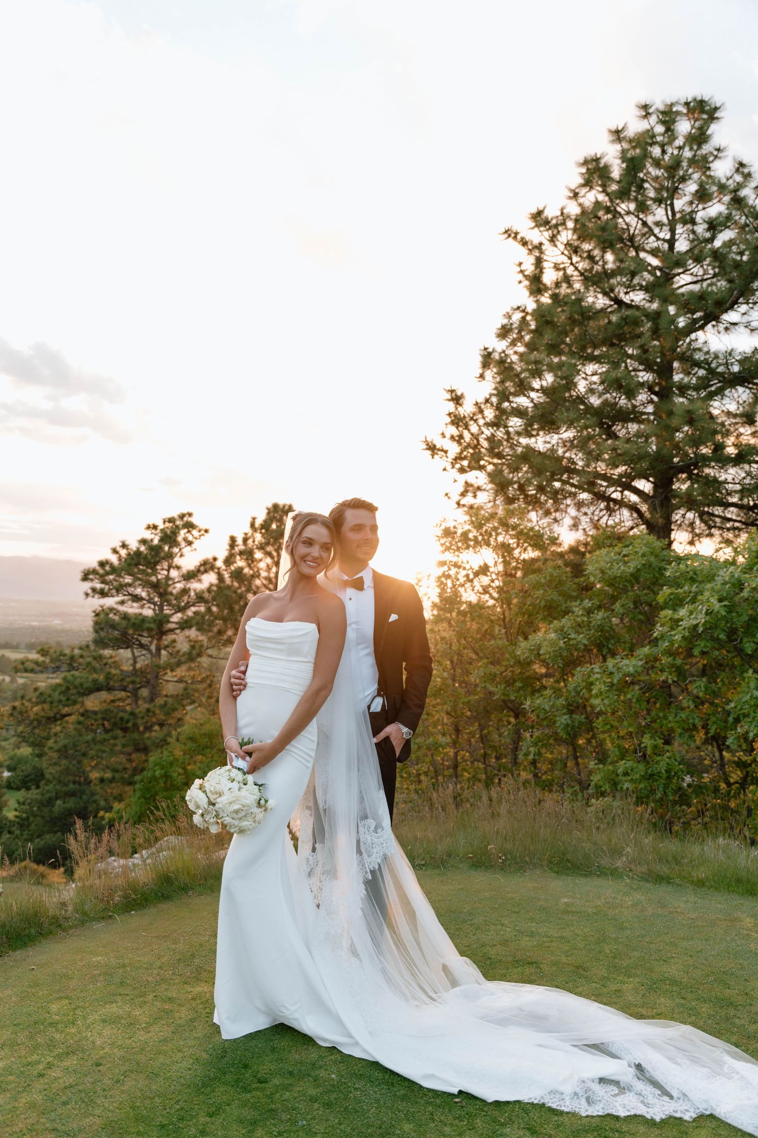 Newlyweds pose together on grassy hillside during golden hour with flowing wedding dress train.