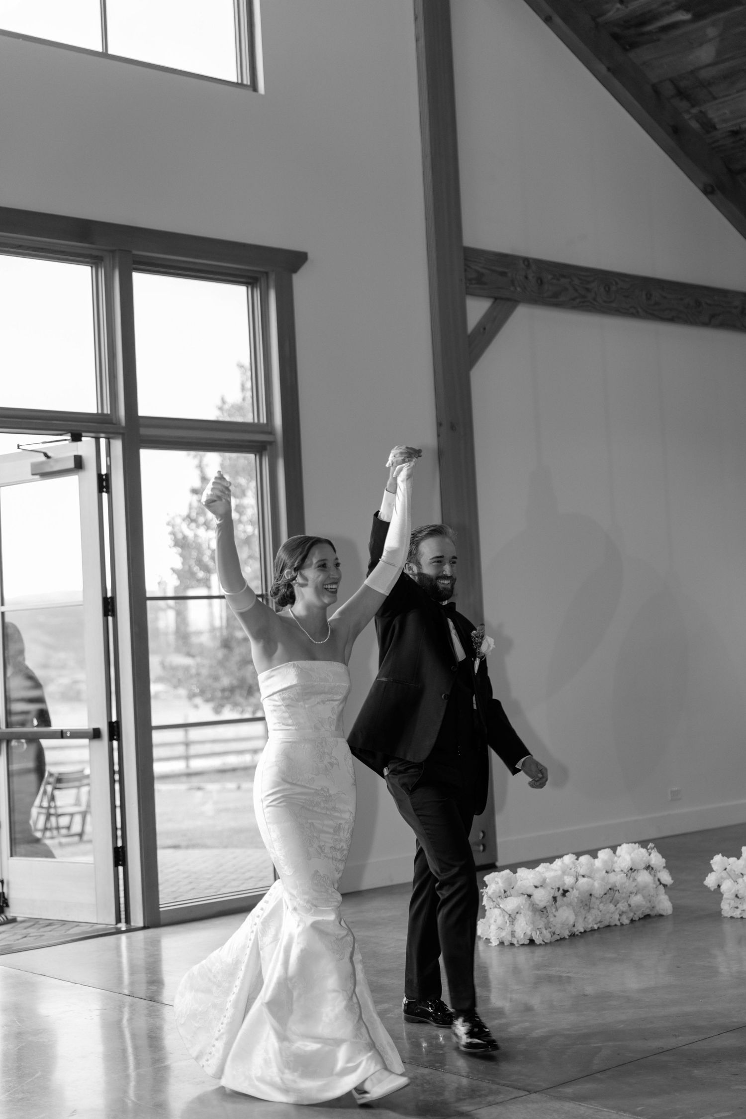 Bride and groom raise hands in celebration as they make grand entrance at wedding reception in black and white photo.