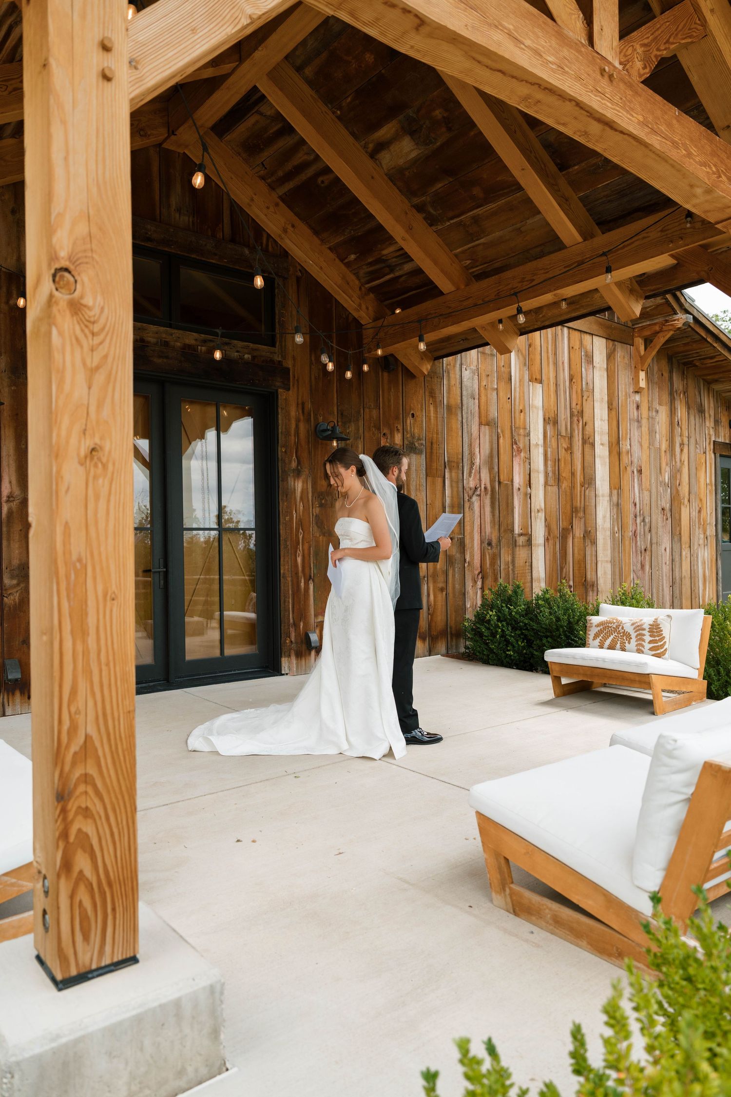 Bride and groom exchange vows on a rustic wooden barn porch with string lights during an outdoor wedding ceremony.
