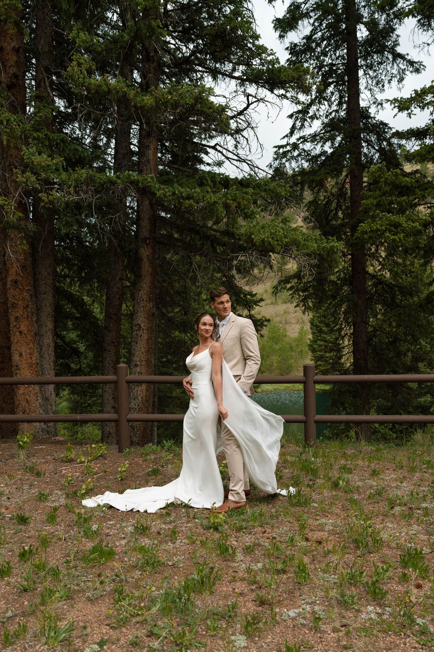 Bride and groom embrace in elegant wedding attire among tall pine trees with mountain backdrop.