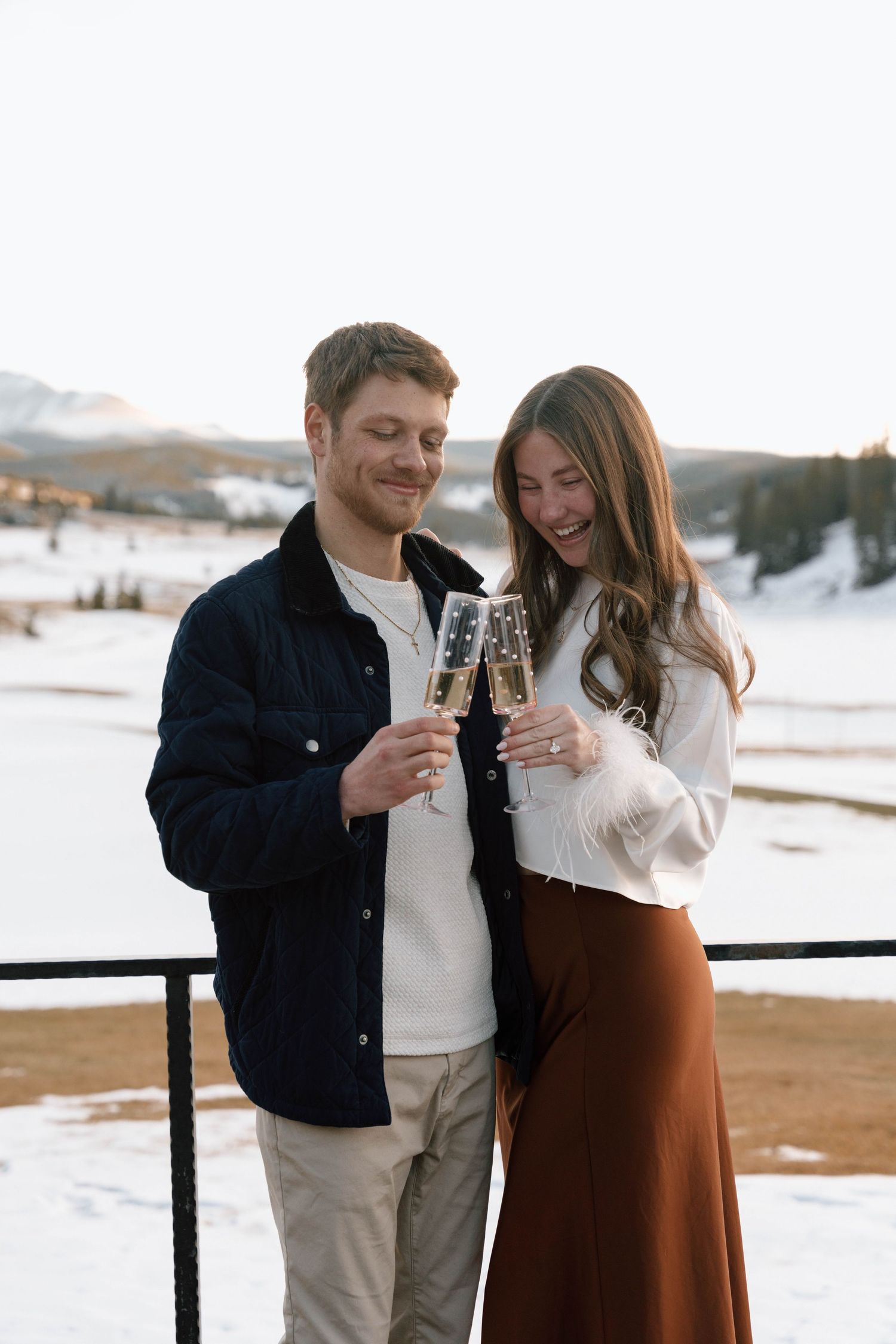 Couple toasting with champagne glasses on a snowy mountain overlook during sunset.