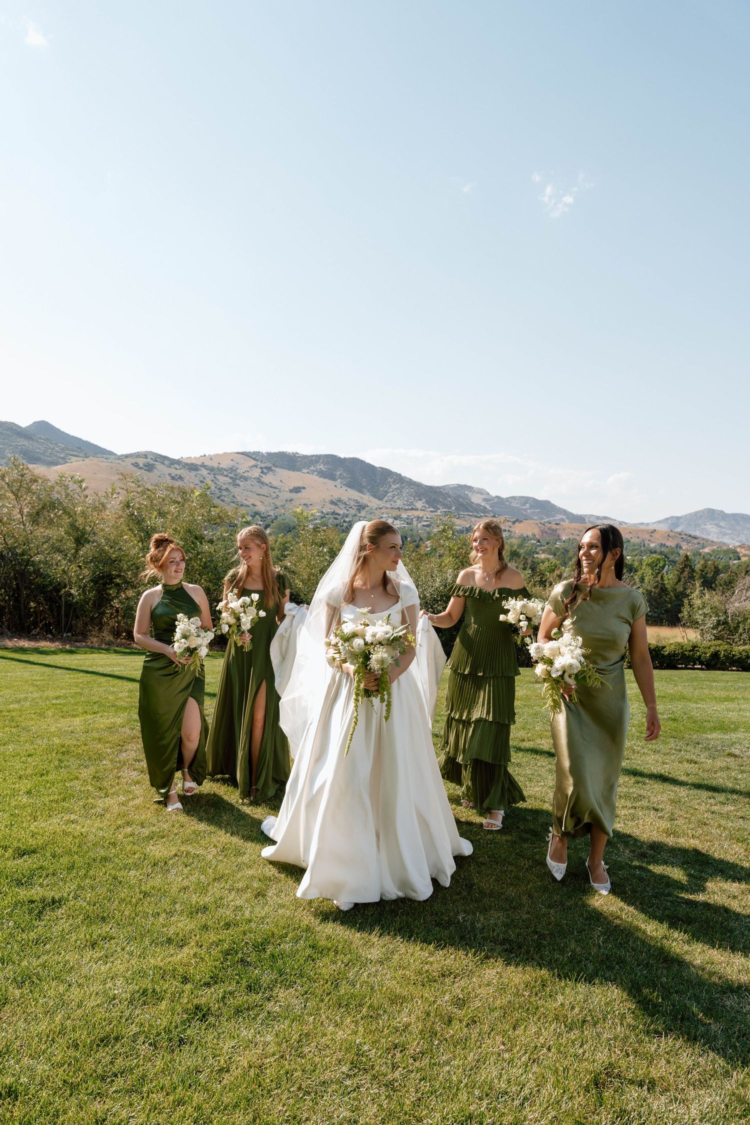Bridal party in sage green dresses walks together across sunny lawn with mountain views.