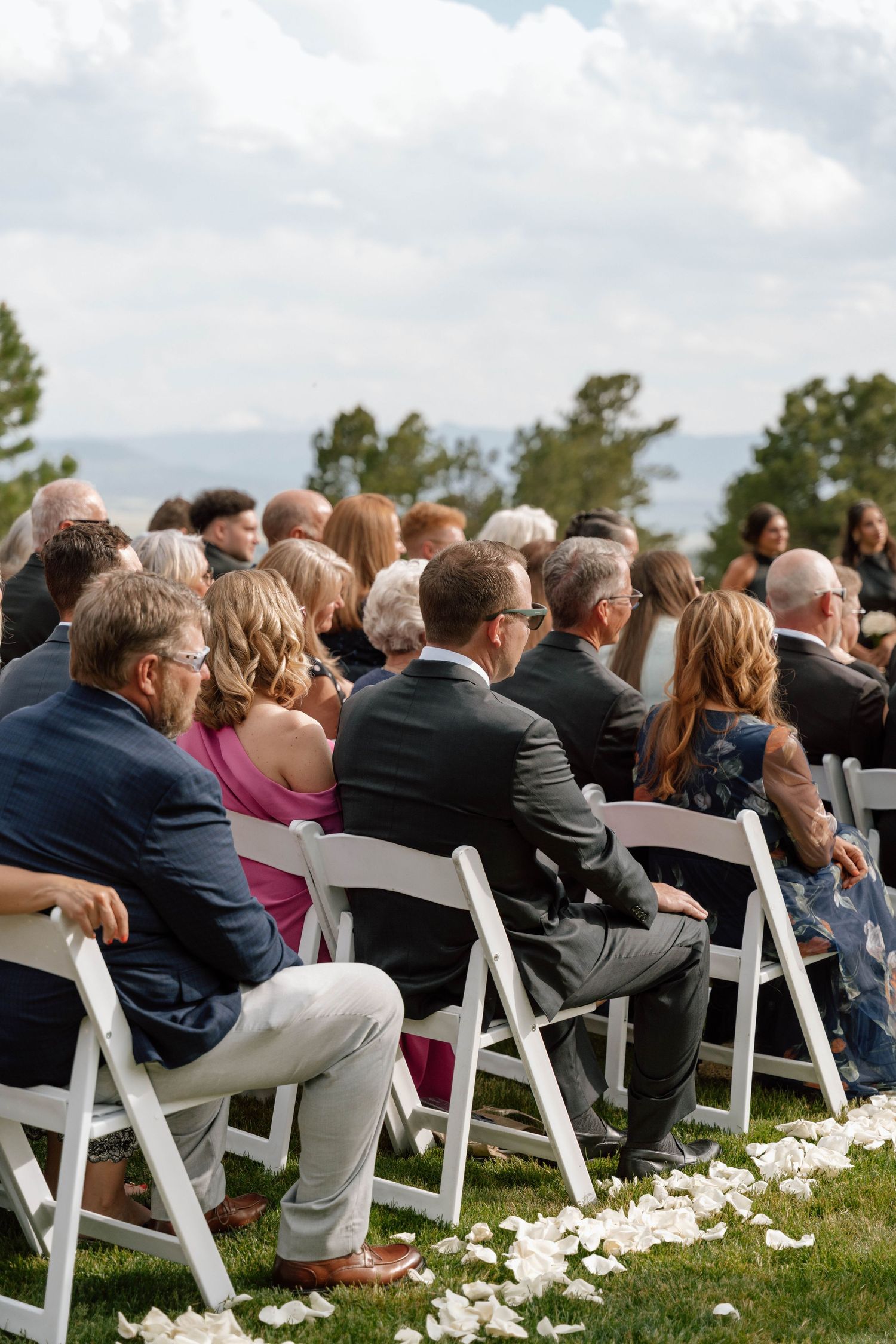 Wedding guests seated in white chairs with flower petals scattered on grass during outdoor ceremony.