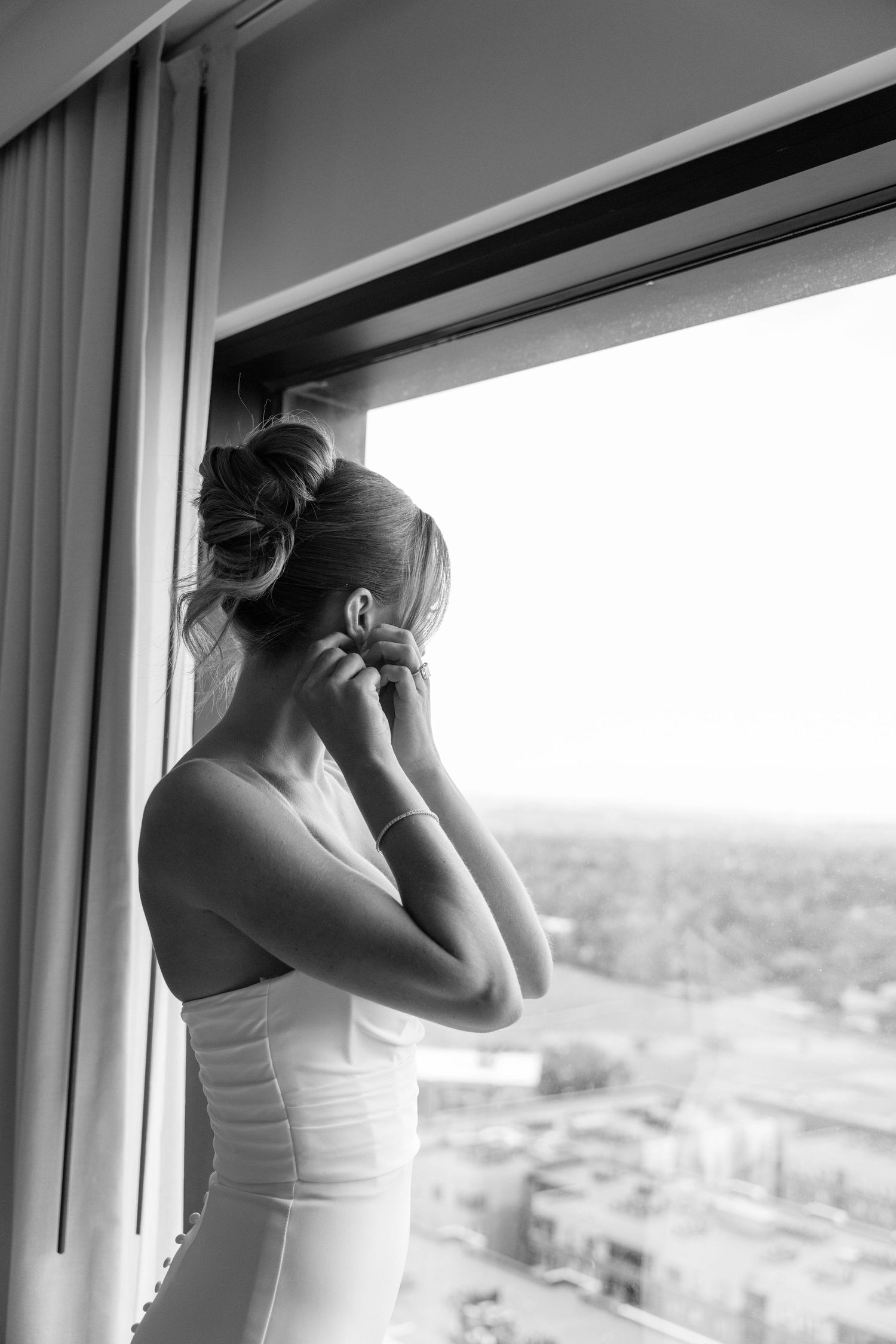 A silhouette in a white dress puts on earrings while standing by a bright window with a city view.