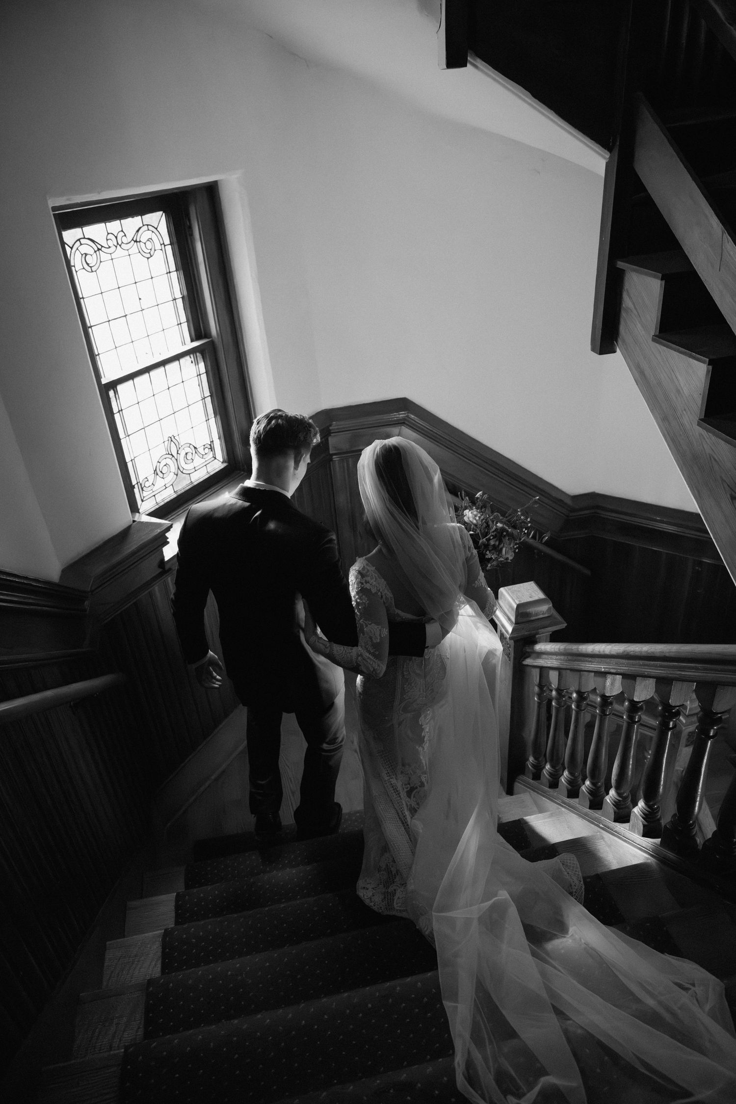 Black and white photo of bride and groom walking up elegant wooden staircase with stained glass window.