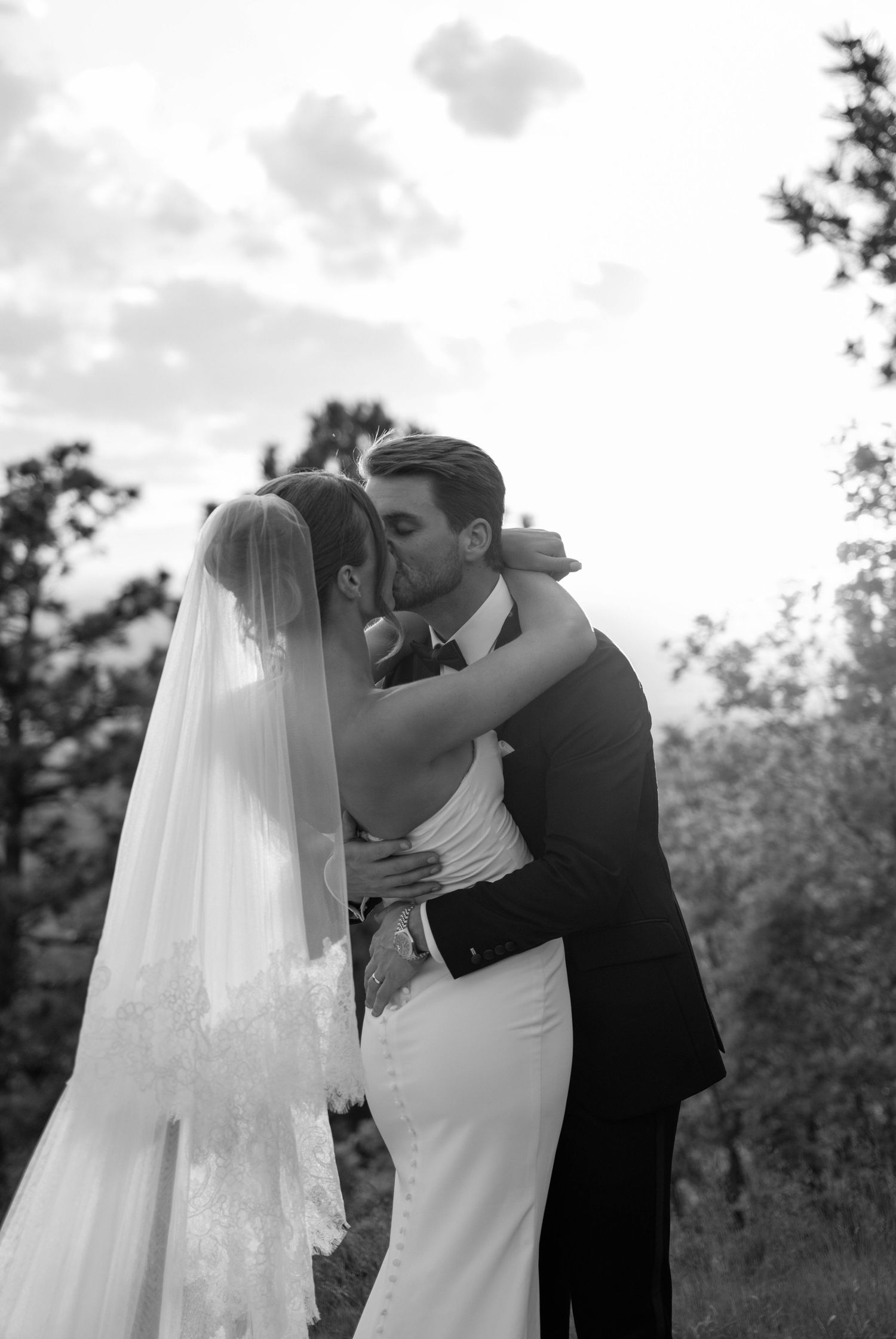 Black and white photo of a bride and groom sharing a romantic kiss outdoors, bride wearing a lace veil and fitted gown.