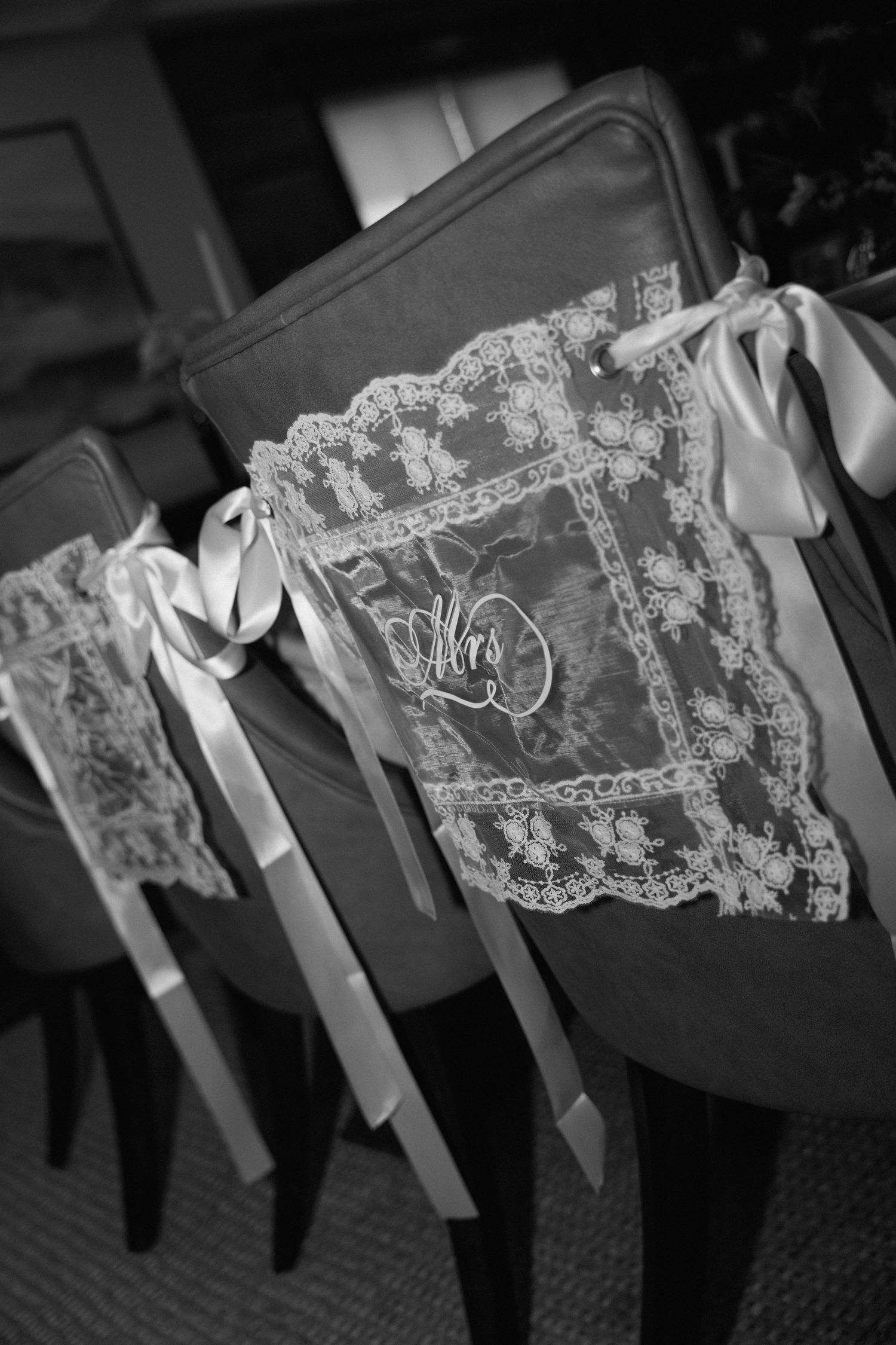 Black and white photo of wedding chairs decorated with lace signs reading Mrs and satin ribbon bows.
