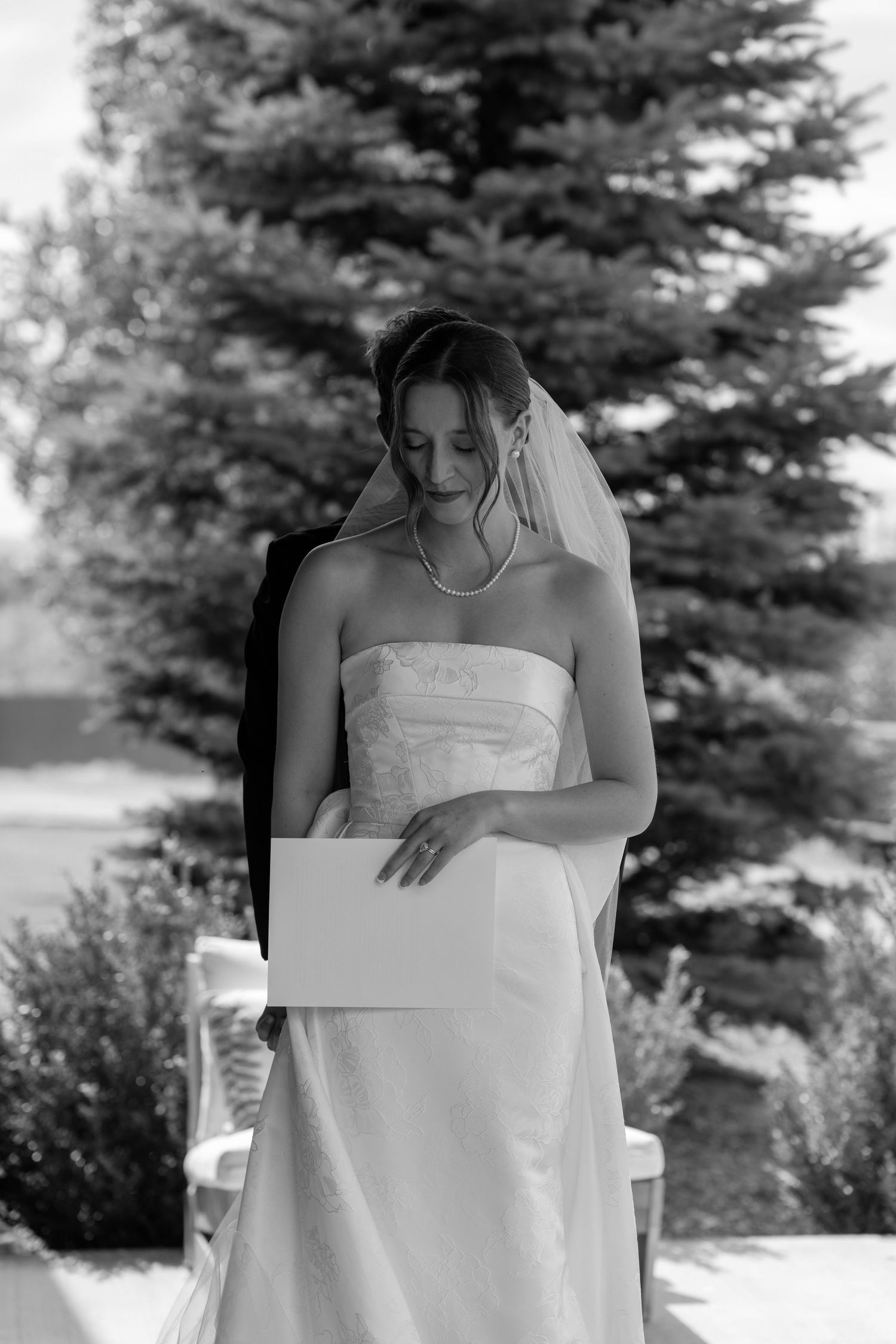 Black and white photo of a bride in a strapless gown and veil reading a card outdoors near evergreen trees.