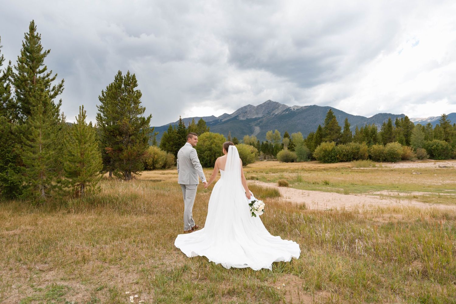 Bride and groom holding hands in a mountain meadow with evergreen trees and dramatic cloudy skies in the background.