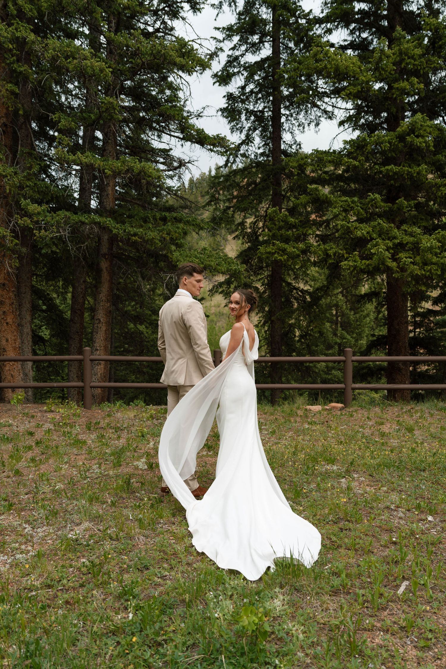 Bride and groom standing together in forest meadow with flowing wedding dress train.