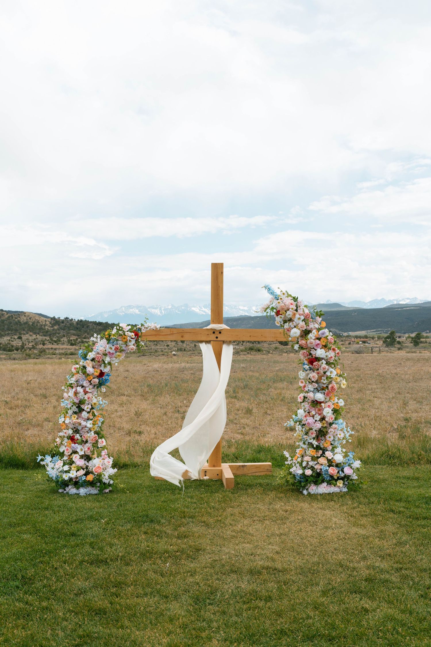 Wooden cross wedding arch adorned with colorful flowers and white fabric in open field.