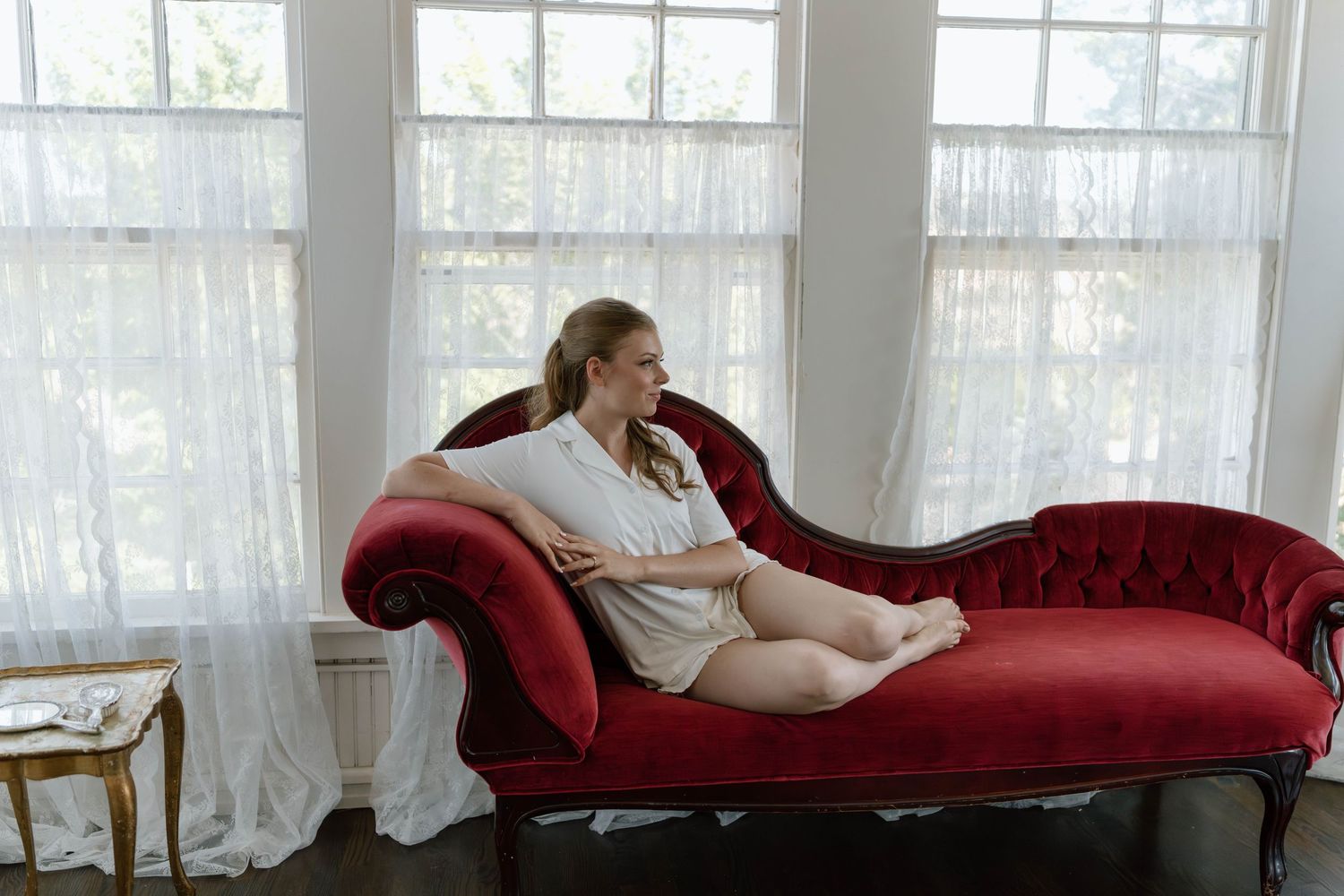 Elegant red velvet chaise lounge positioned in a bright room with white sheer curtains.