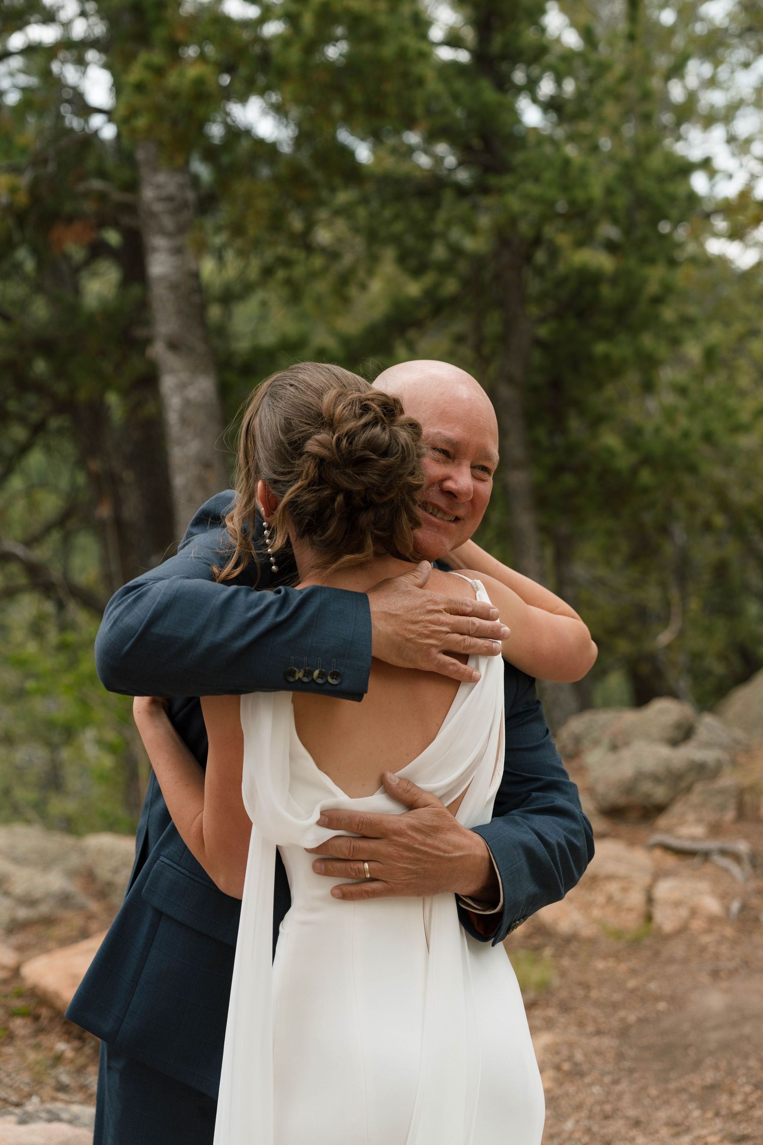 A bald man in a navy suit smiles while embracing a bride in a white backless gown outdoors.