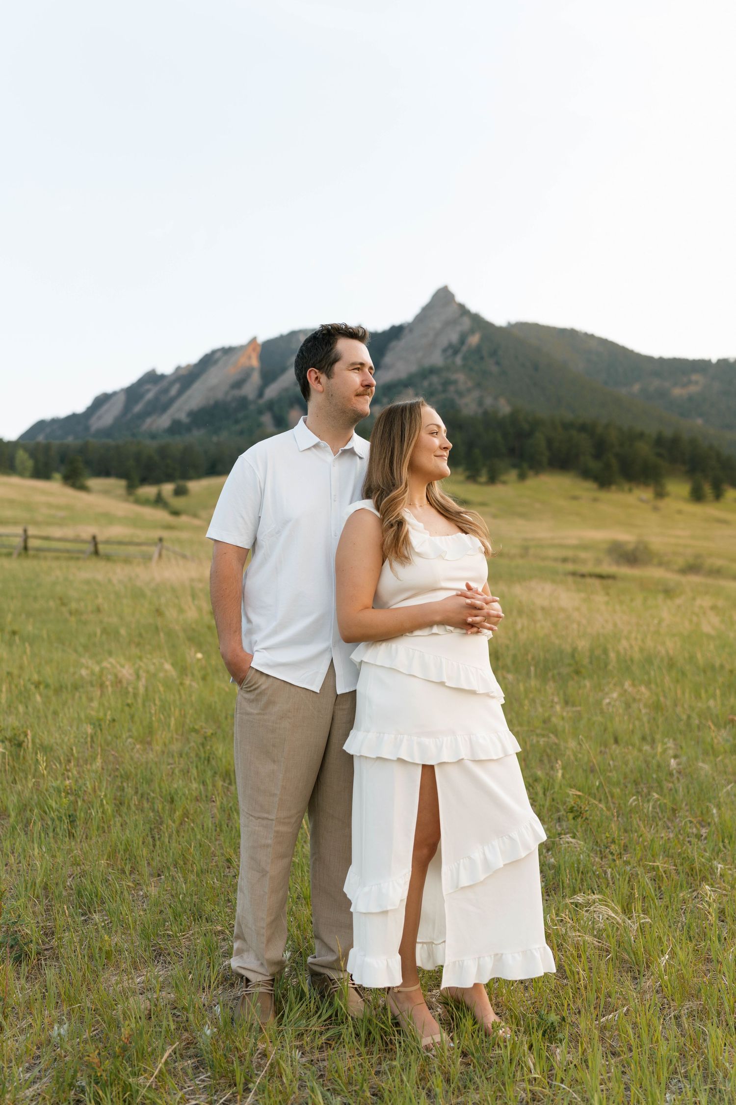Couple in white outfits standing in green meadow with Flatirons mountain backdrop in Boulder, Colorado.