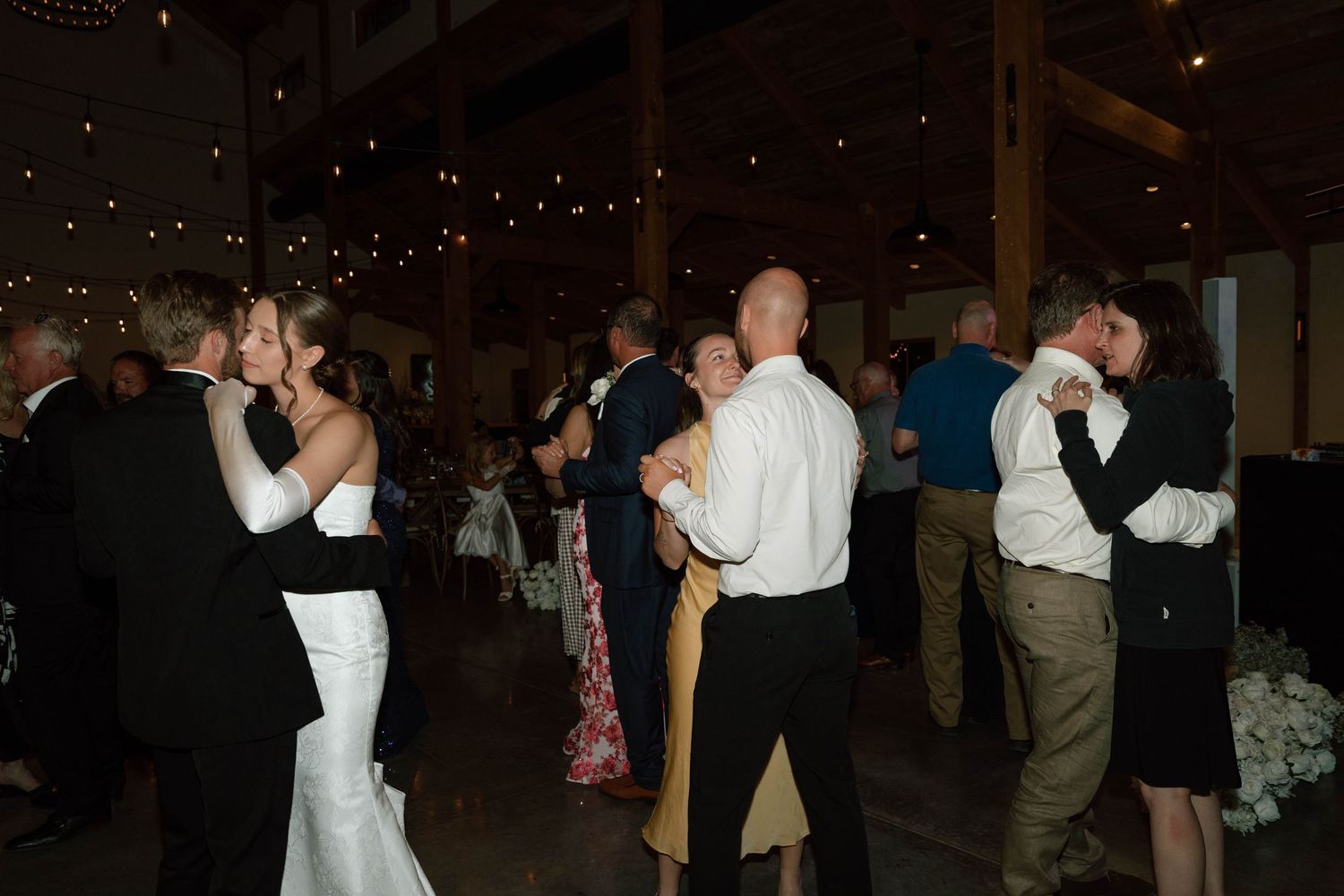 Couples share romantic dances on a dimly lit barn wedding reception dance floor adorned with string lights overhead.