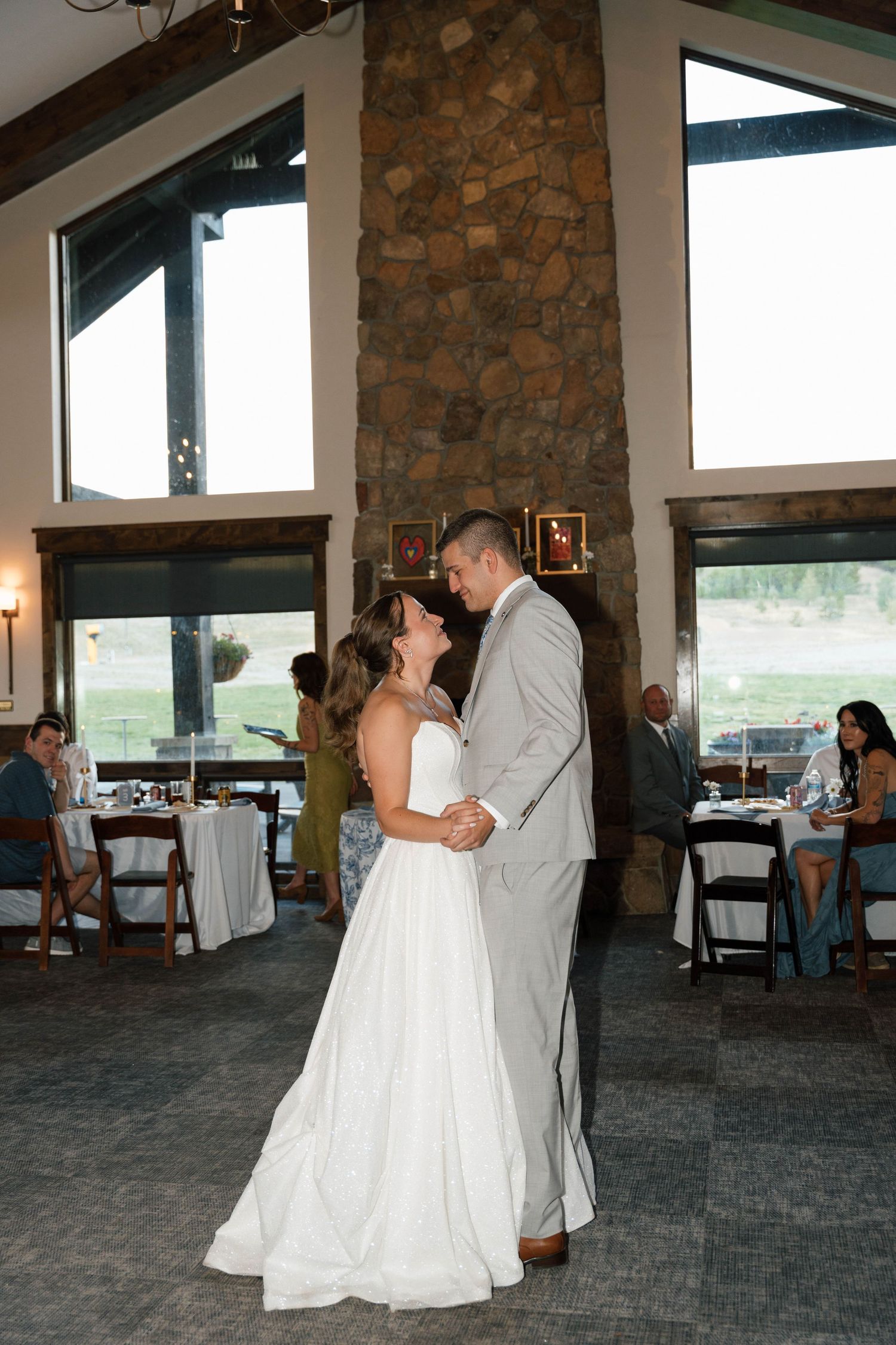 Bride and groom share their first dance in an elegant venue with a stone fireplace and large windows.