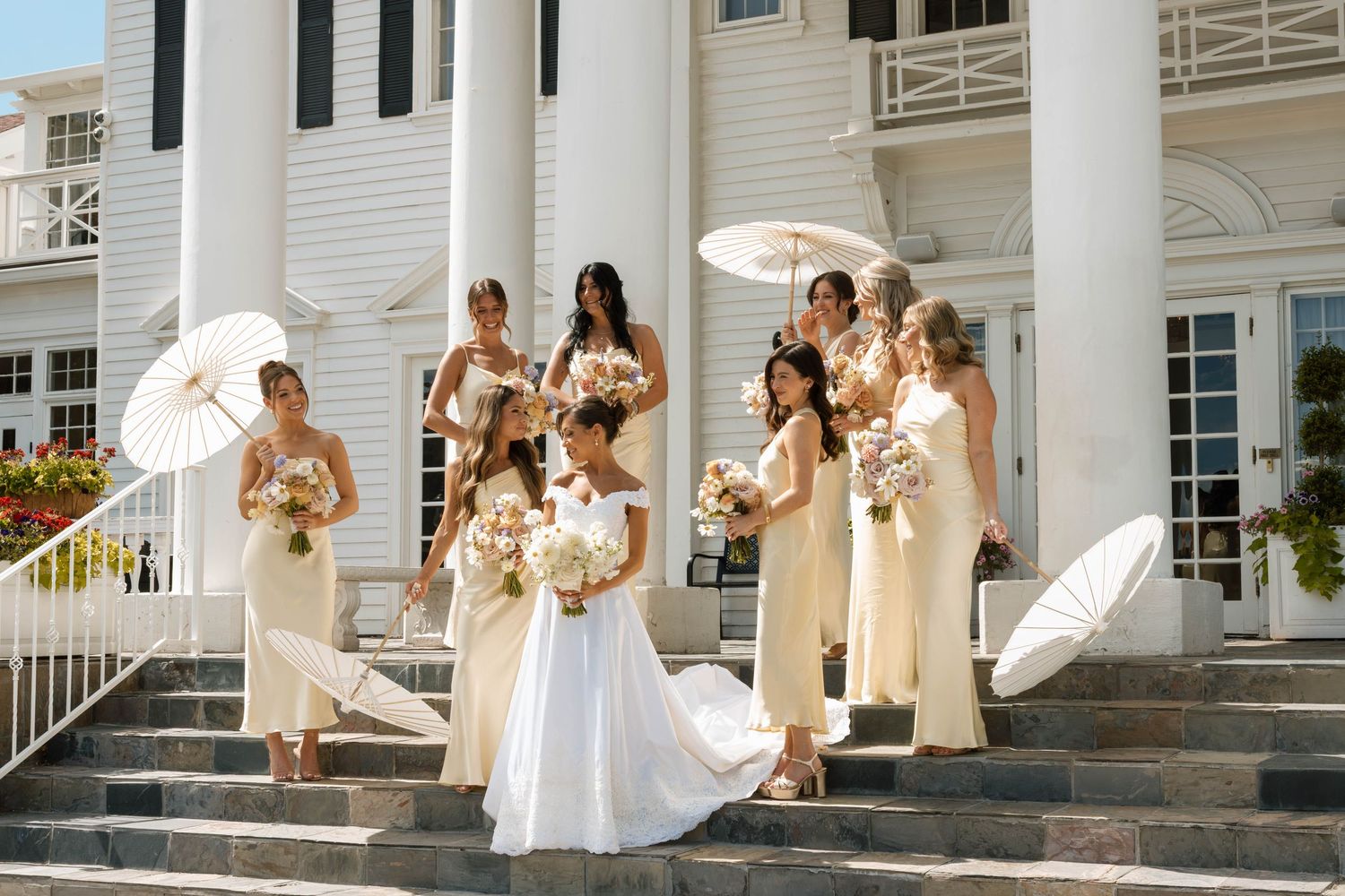 Bride in white gown with bridesmaids in cream dresses holding parasols and bouquets on steps of elegant white mansion.