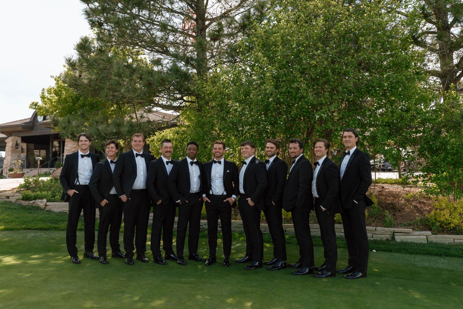 A group in formal black tuxedos posing on a green lawn with pine trees in the background.