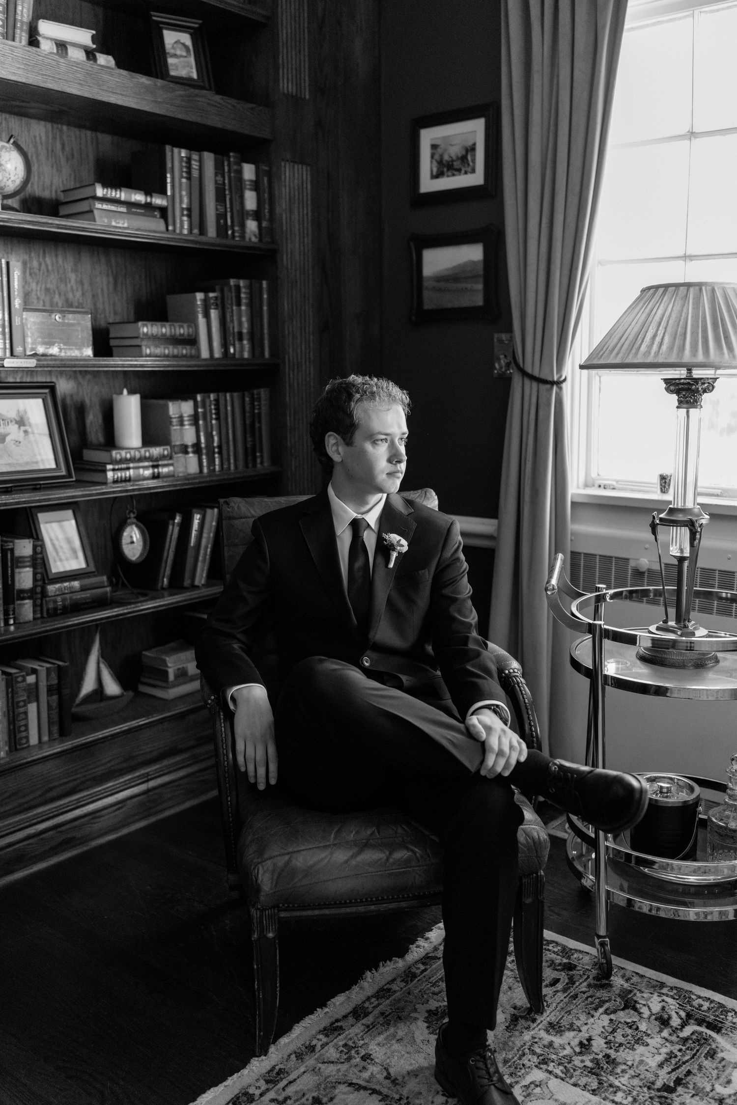 A groom in a dark suit sits in a leather chair in an elegant library, gazing toward a window in this black and white photo.