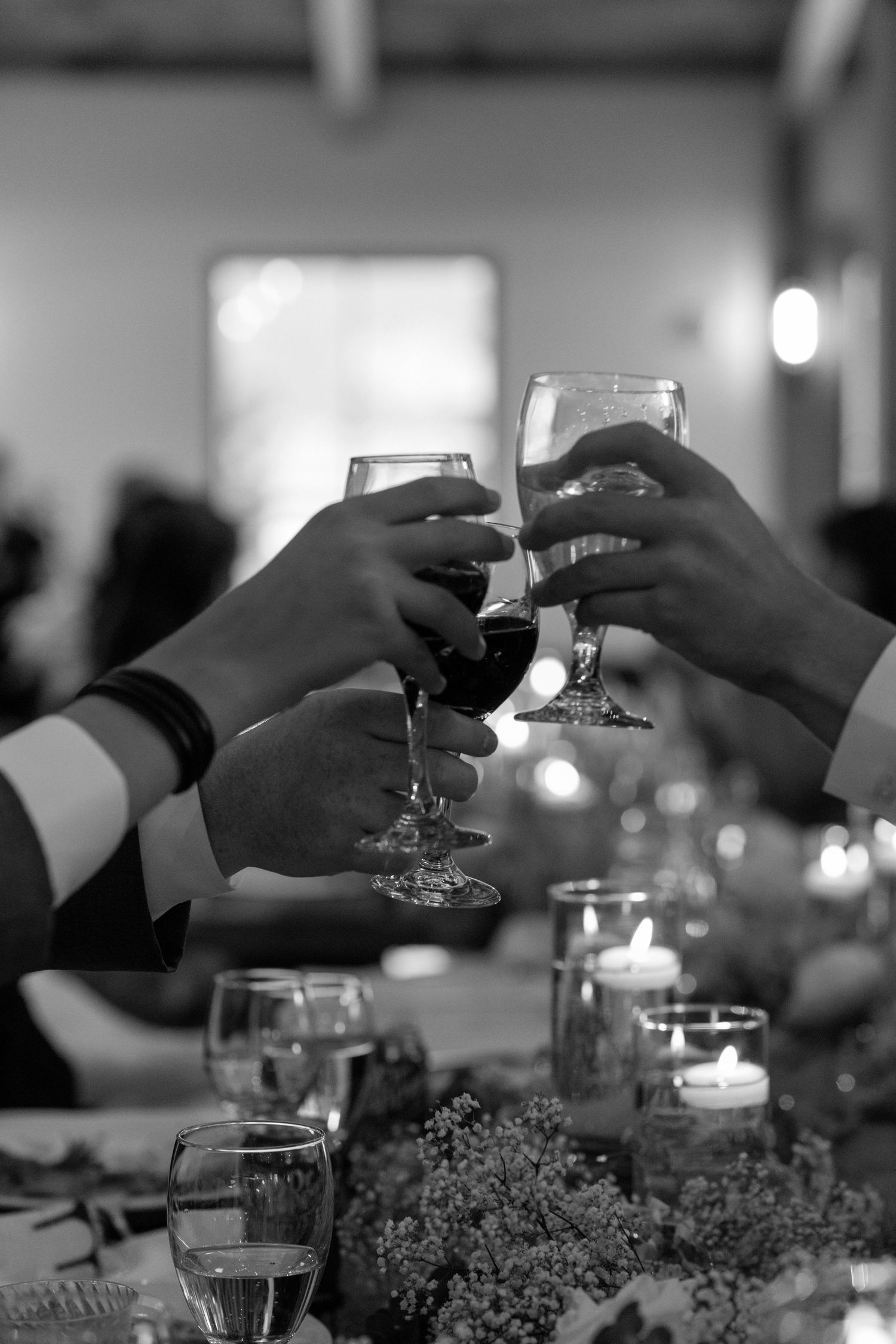 Black and white photo of hands clinking wine glasses at a candlelit celebration dinner table.