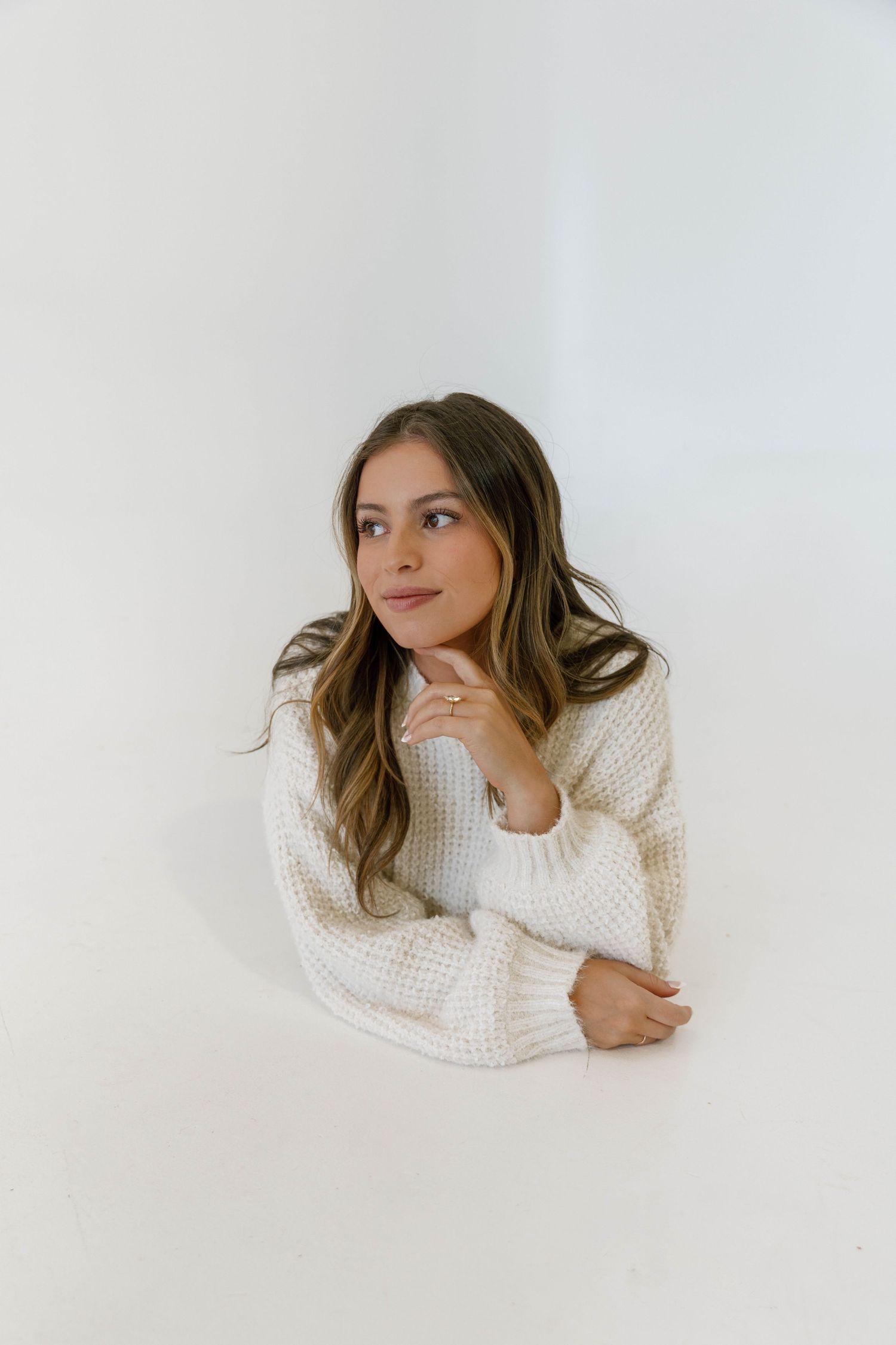 Woman in cozy cream knit sweater poses thoughtfully against white background.