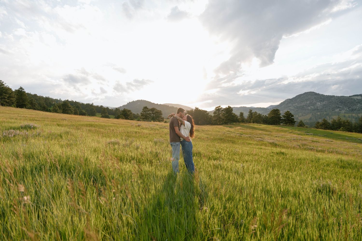 A couple embracing in a vast mountain meadow with dramatic cloudy skies overhead.