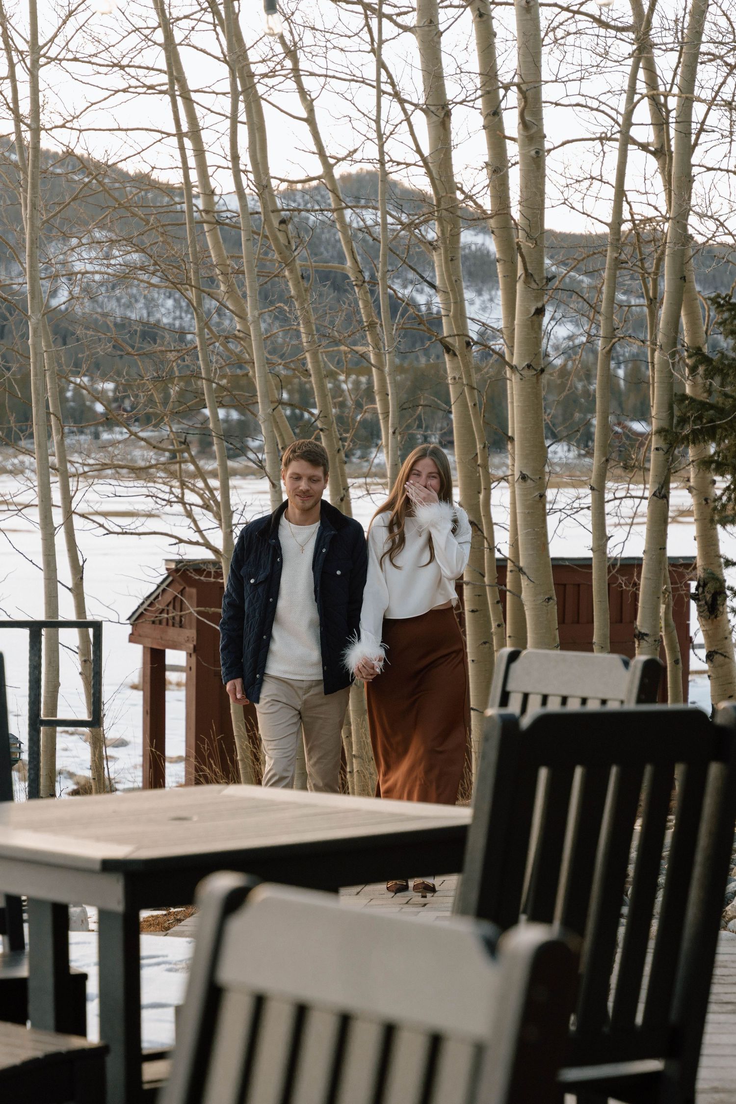 Couple walking on snow-covered deck with aspen trees and mountain landscape in background.