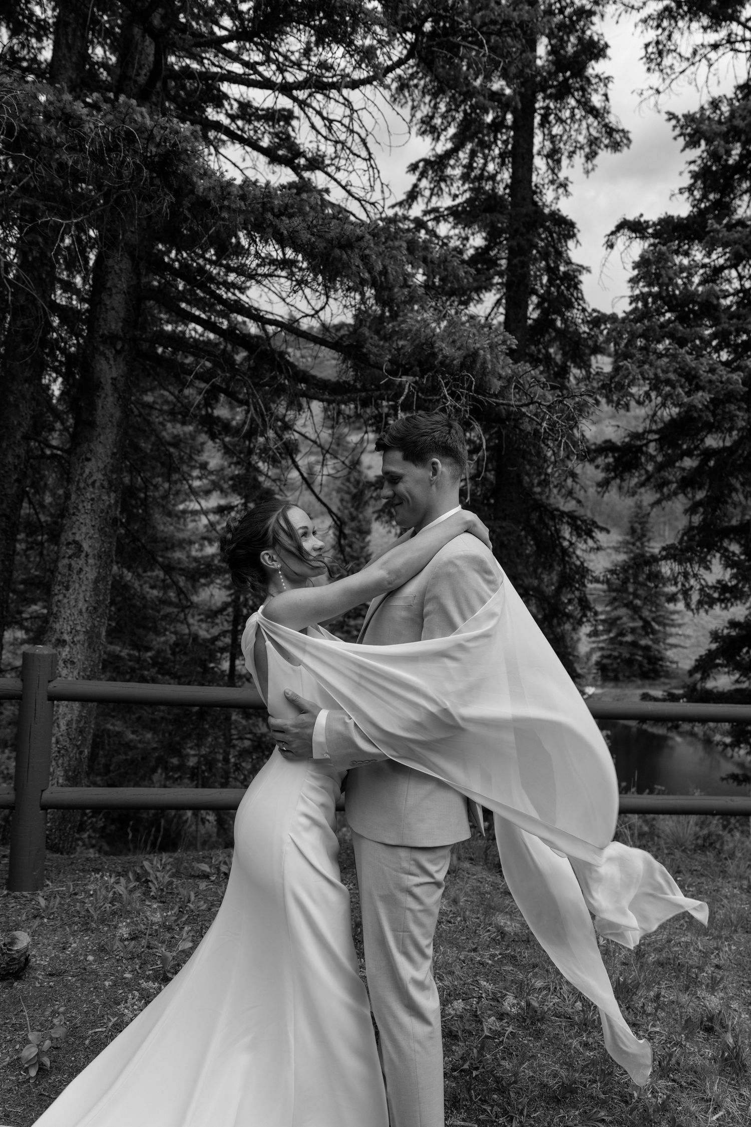 Groom lifts smiling bride in white gown among tall pine trees near a mountain lake.