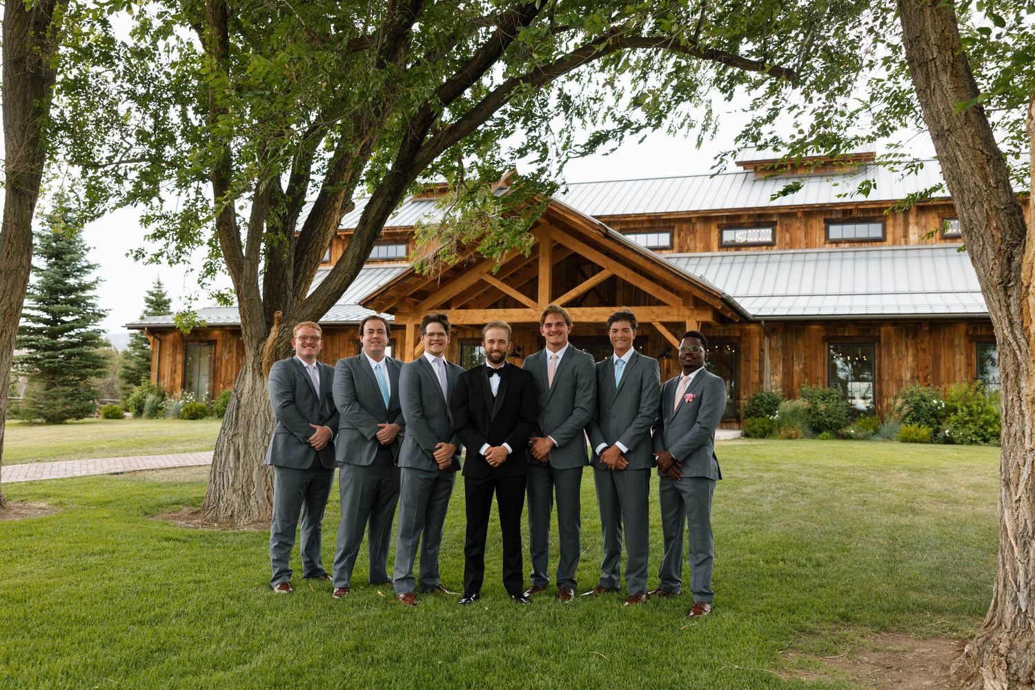 Groom and groomsmen in gray suits pose outdoors under trees near a rustic wooden barn venue.