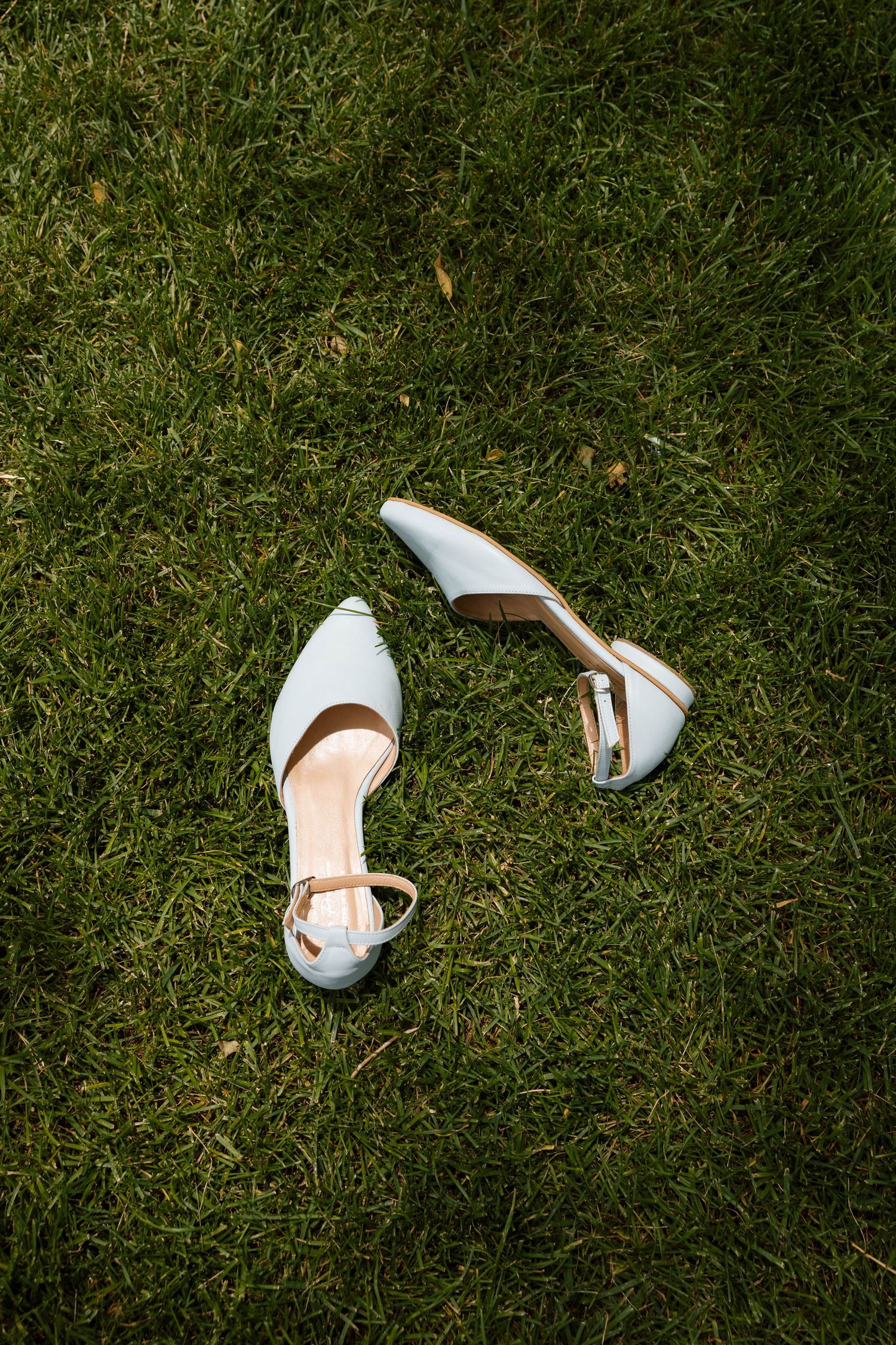 White bridal heels with ankle straps resting on lush green grass, perfect for an outdoor wedding ceremony.