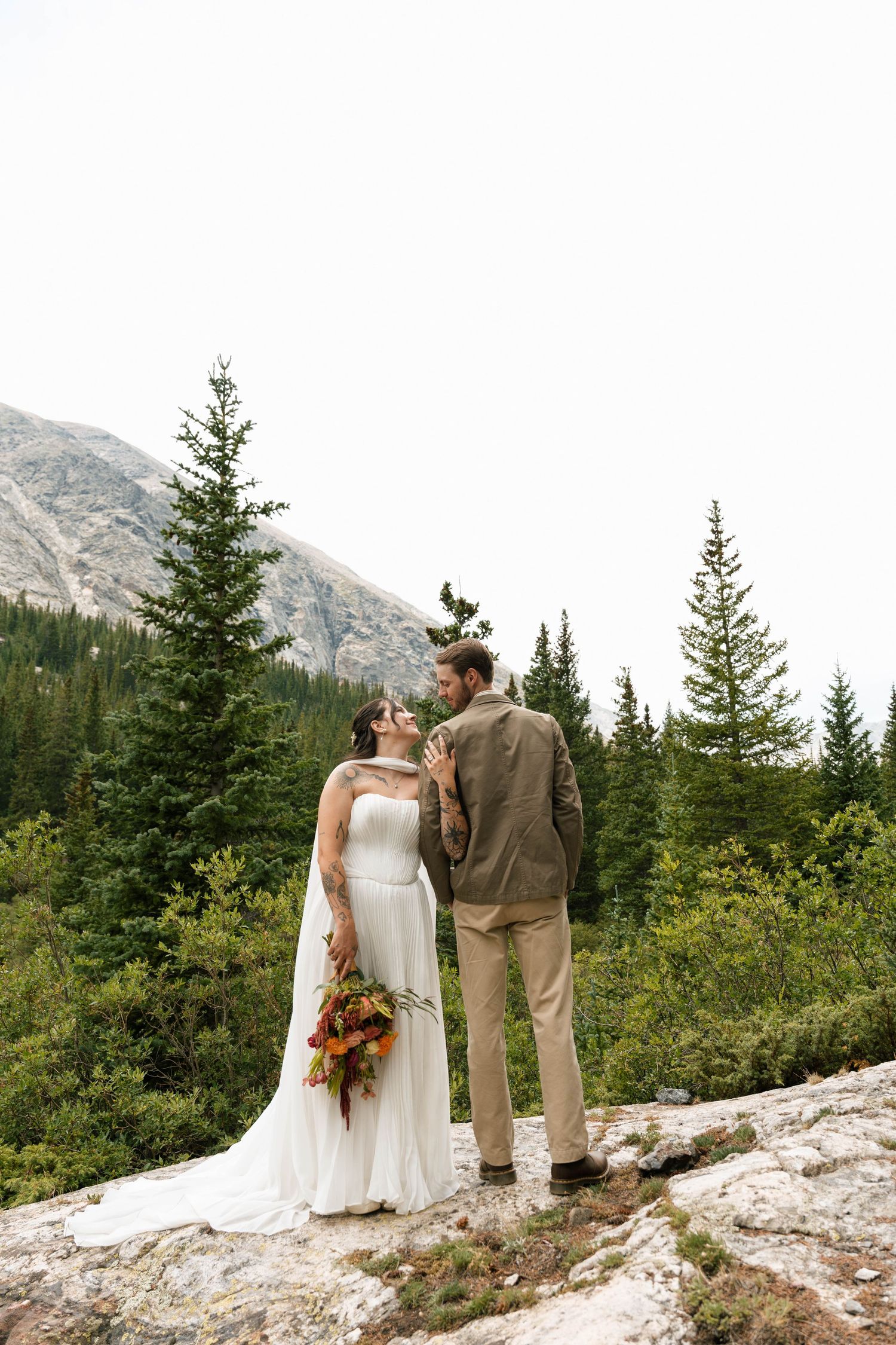 Bride and groom stand on rocky mountain overlook surrounded by evergreen trees, bride holding autumn bouquet in white gown.
