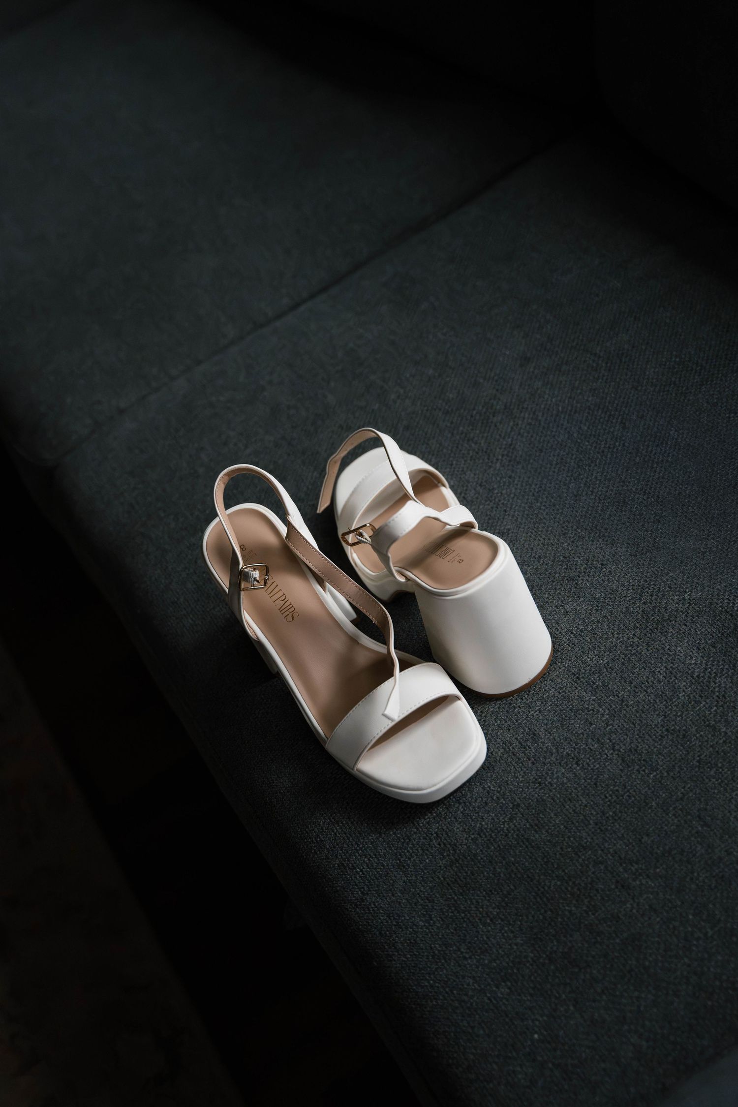 White strappy block heel sandals with ankle strap placed on dark gray upholstered surface.