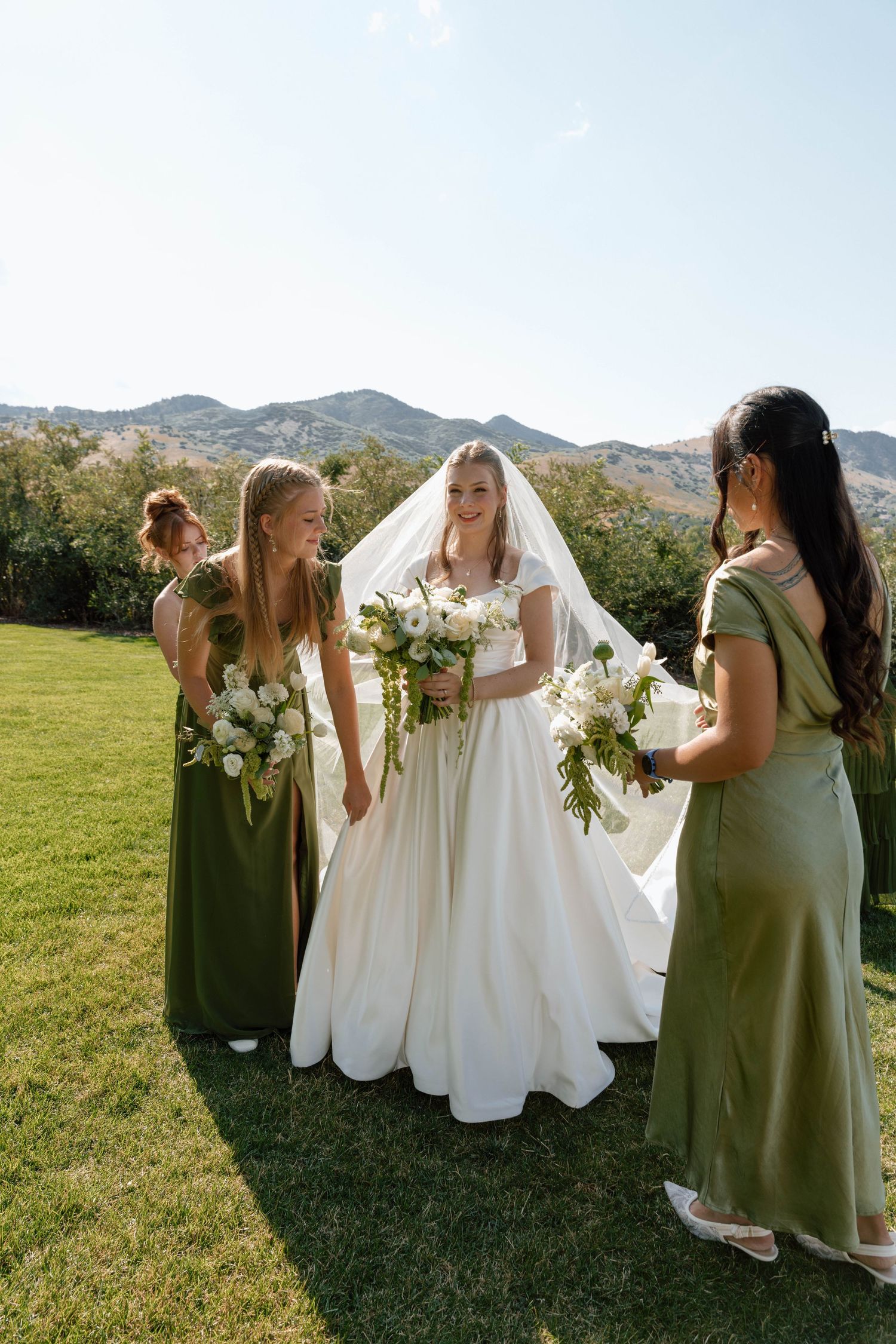 Wedding party in sage green dresses gathers on grass with mountain backdrop.