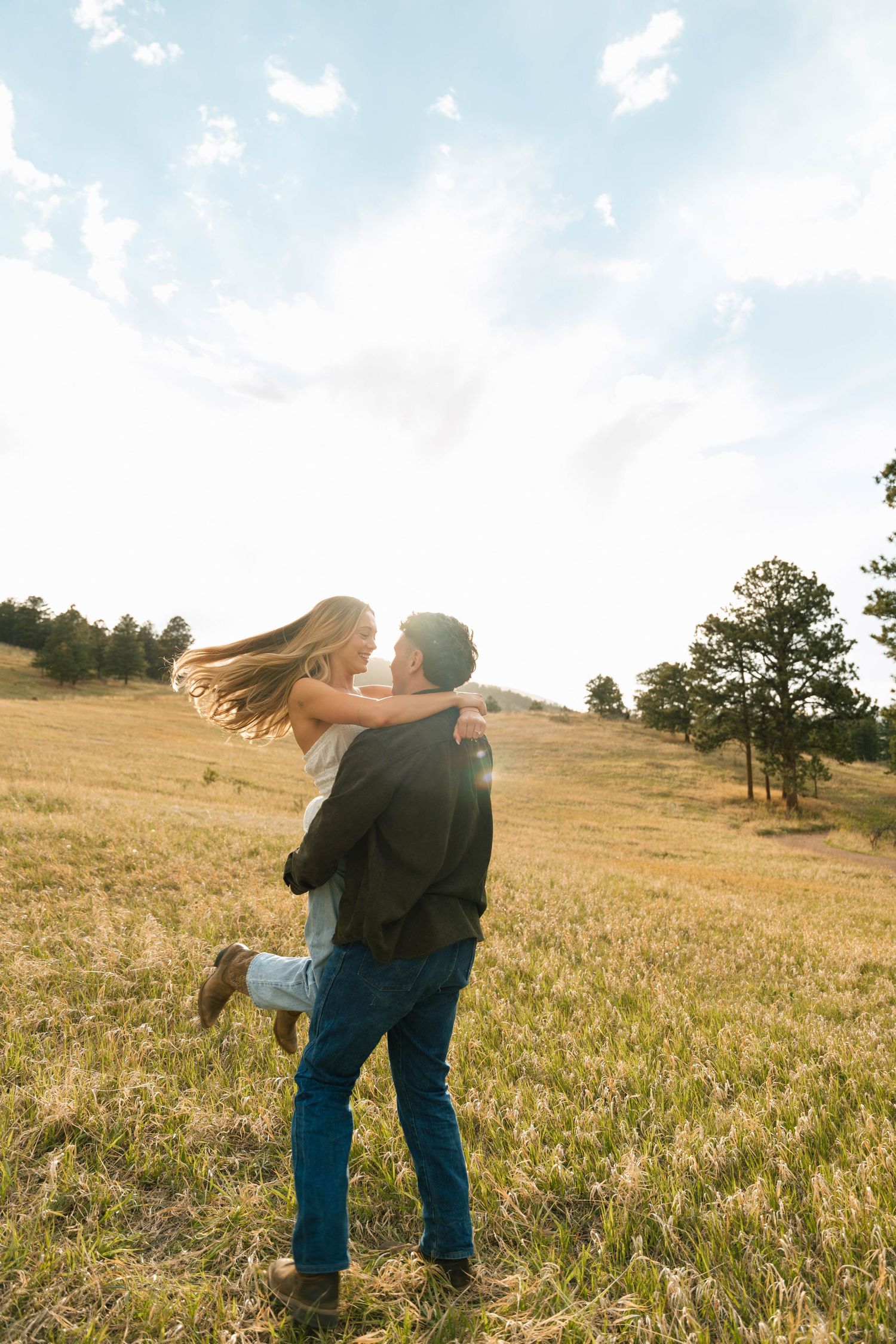 A couple embraces in a sunlit golden field, the man lifting the woman as her hair flows in the breeze near pine trees.
