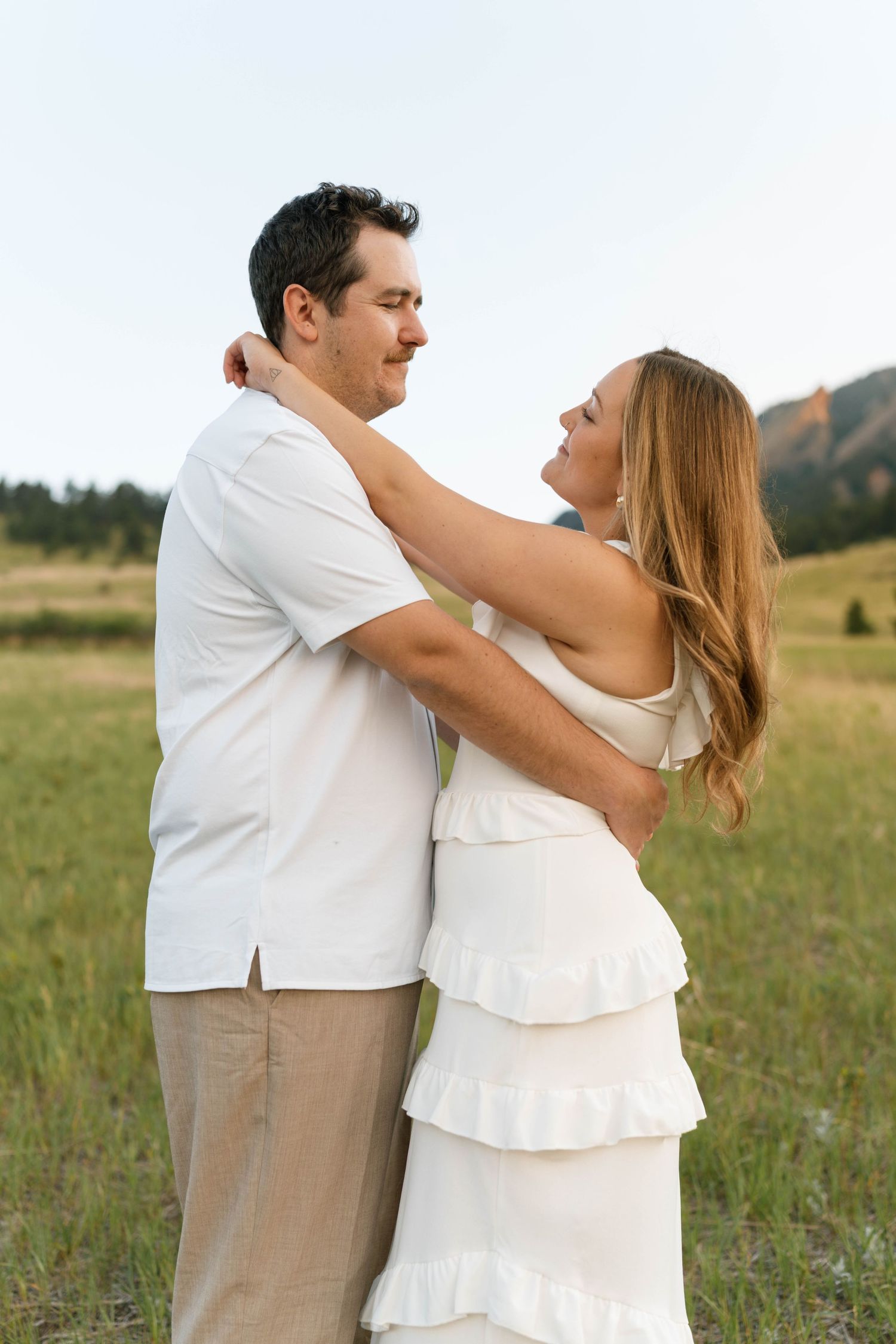 Couple embracing in a green meadow, woman in white ruffled dress with arms around mans neck, mountains behind.