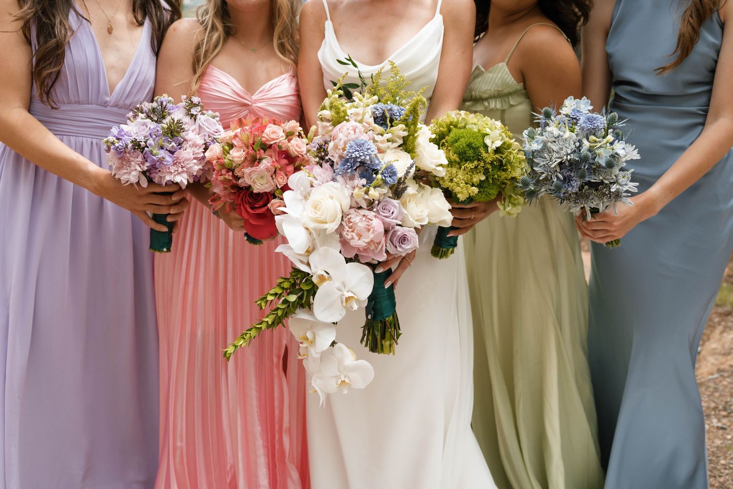 Bride and bridesmaids holding colorful floral bouquets in pastel gowns of lavender, pink, green, and blue.